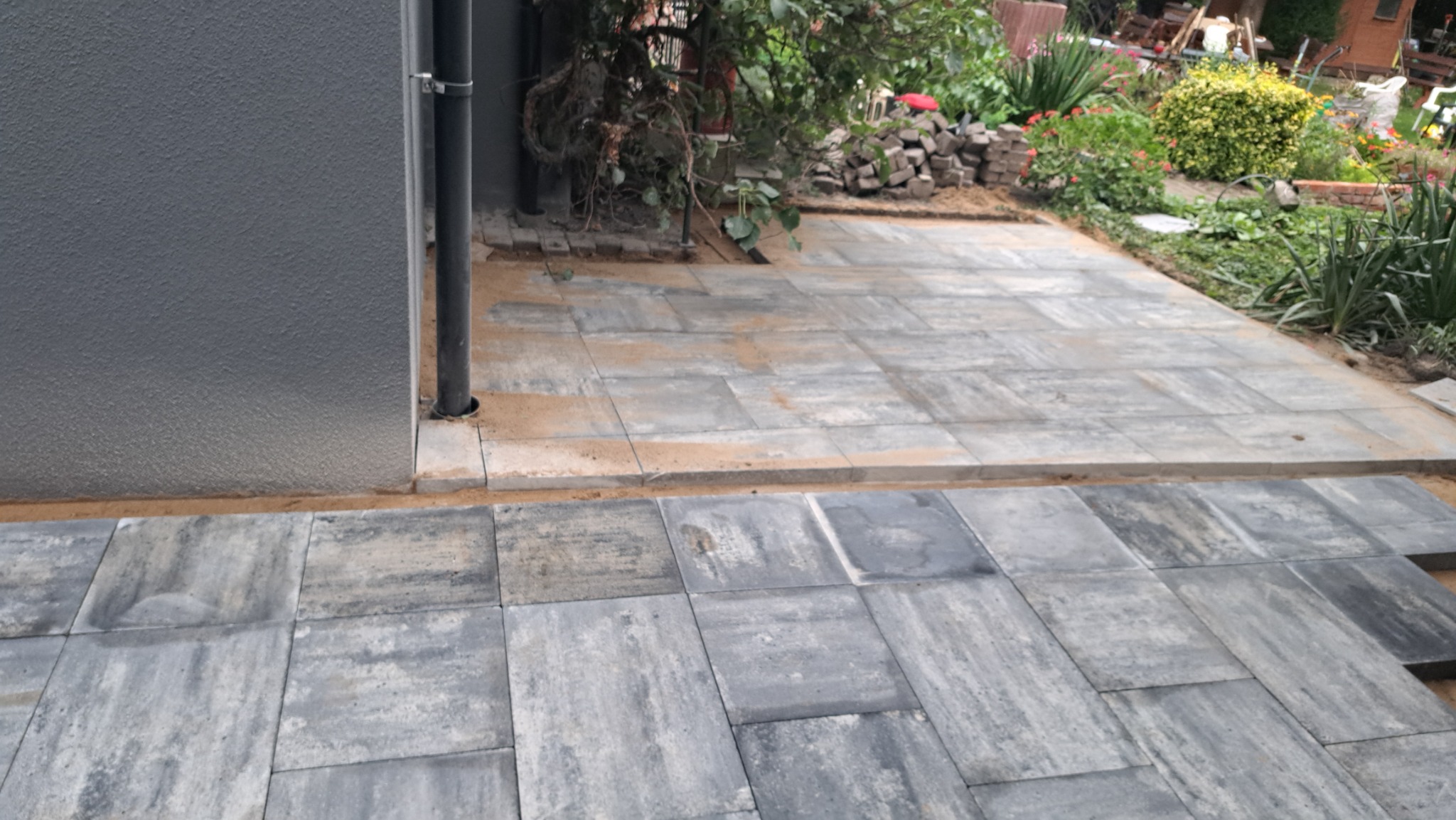 Układanie szarej kostki brukowej na podsypce piaskowej przy ścianie budynku, widoczne niedokończone fragmenty i przygotowane kostki.