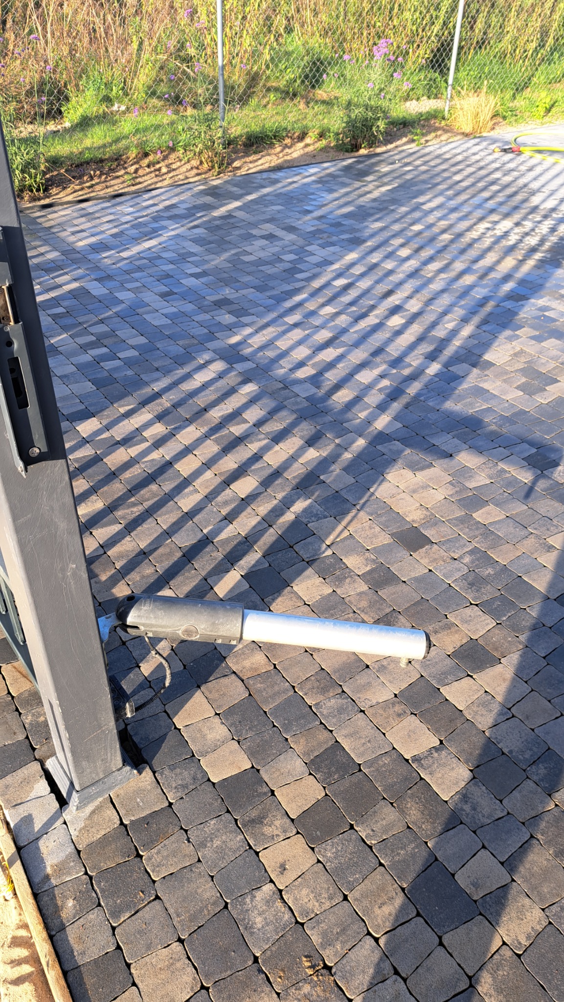 Szara kostka brukowa ułożona w regularny wzór, z widocznym siłownikiem bramy automatycznej i cieniem rzucanym przez ogrodzenie.