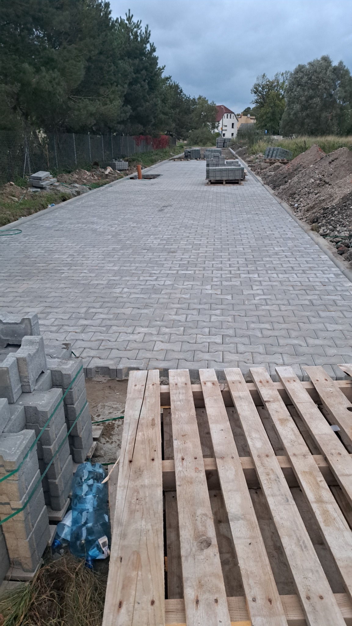 Ułożona szara kostka brukowa typu I na długim podjeździe, paleta z kostką i butelki wody w pobliżu, widok na dom i drzewa w tle, pochmurne niebo.