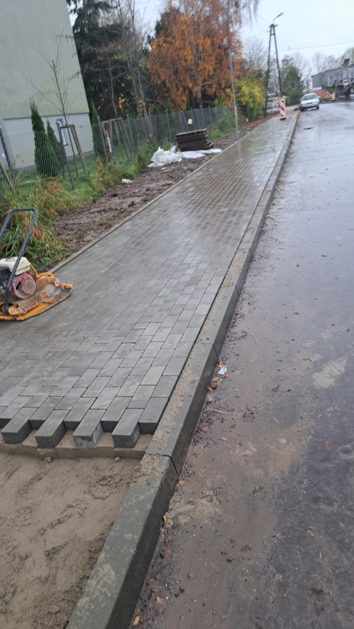 Ułożona część chodnika z szarej kostki, w tle wibro ubijak i ulica po deszczu. Fragment chodnika jeszcze nieukończony, widoczny piasek pod kostką.