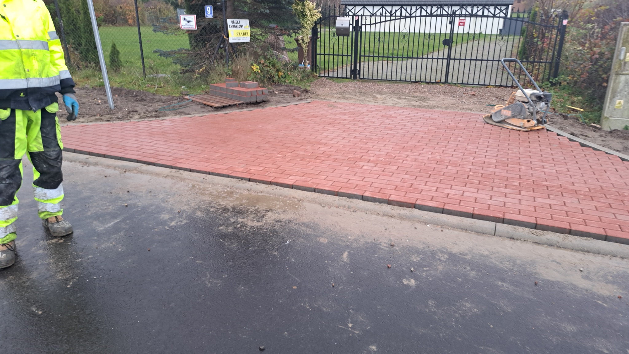 Częściowo ułożona czerwona kostka brukowa przed posesją z czarną bramą, widoczna ubijarka oraz pracownik w odblaskowej odzieży roboczej.