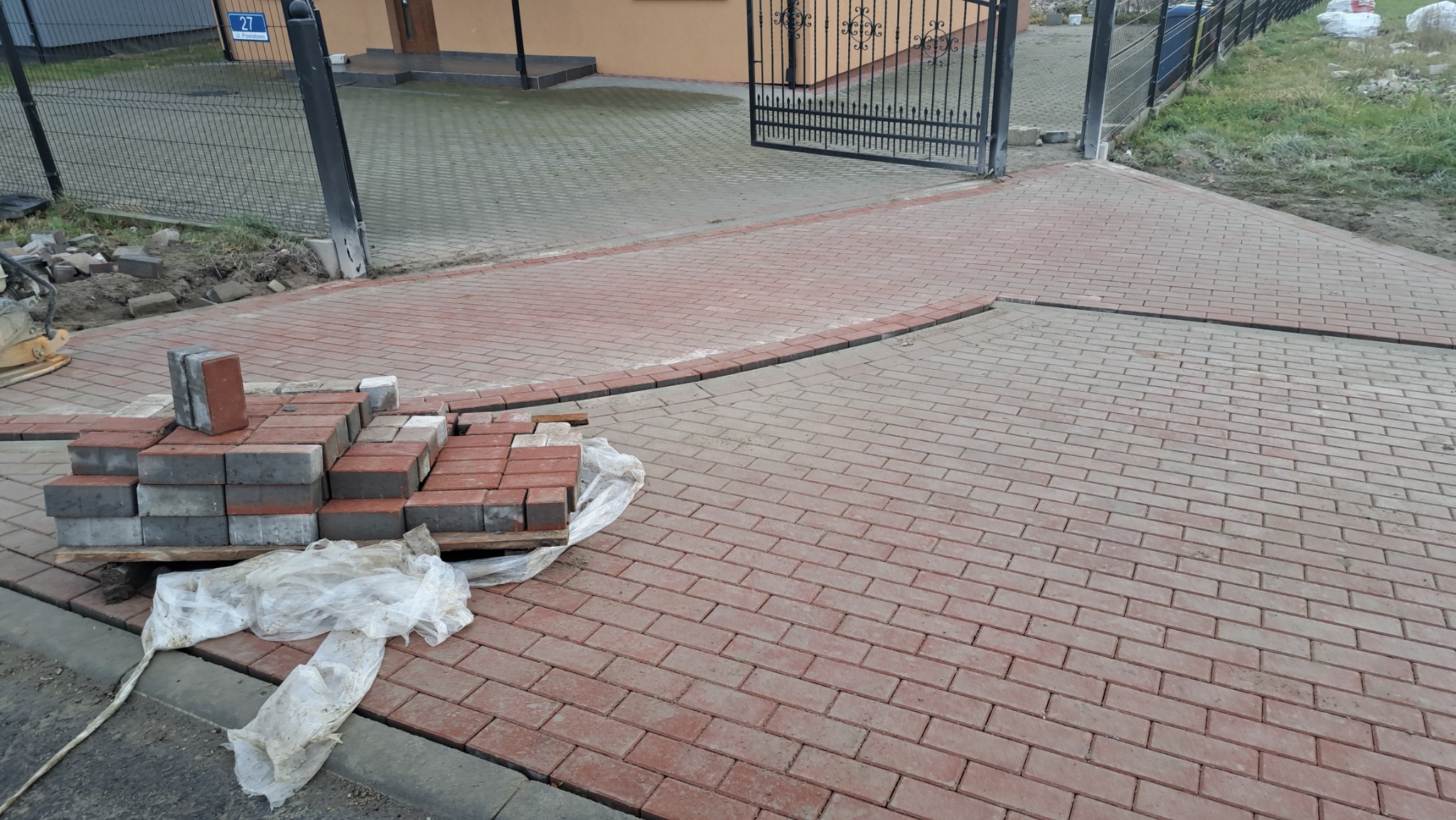 Częściowo ułożona czerwona kostka brukowa na podjeździe, paleta z zapasem kostki, widoczna brama wjazdowa i fragment budynku.