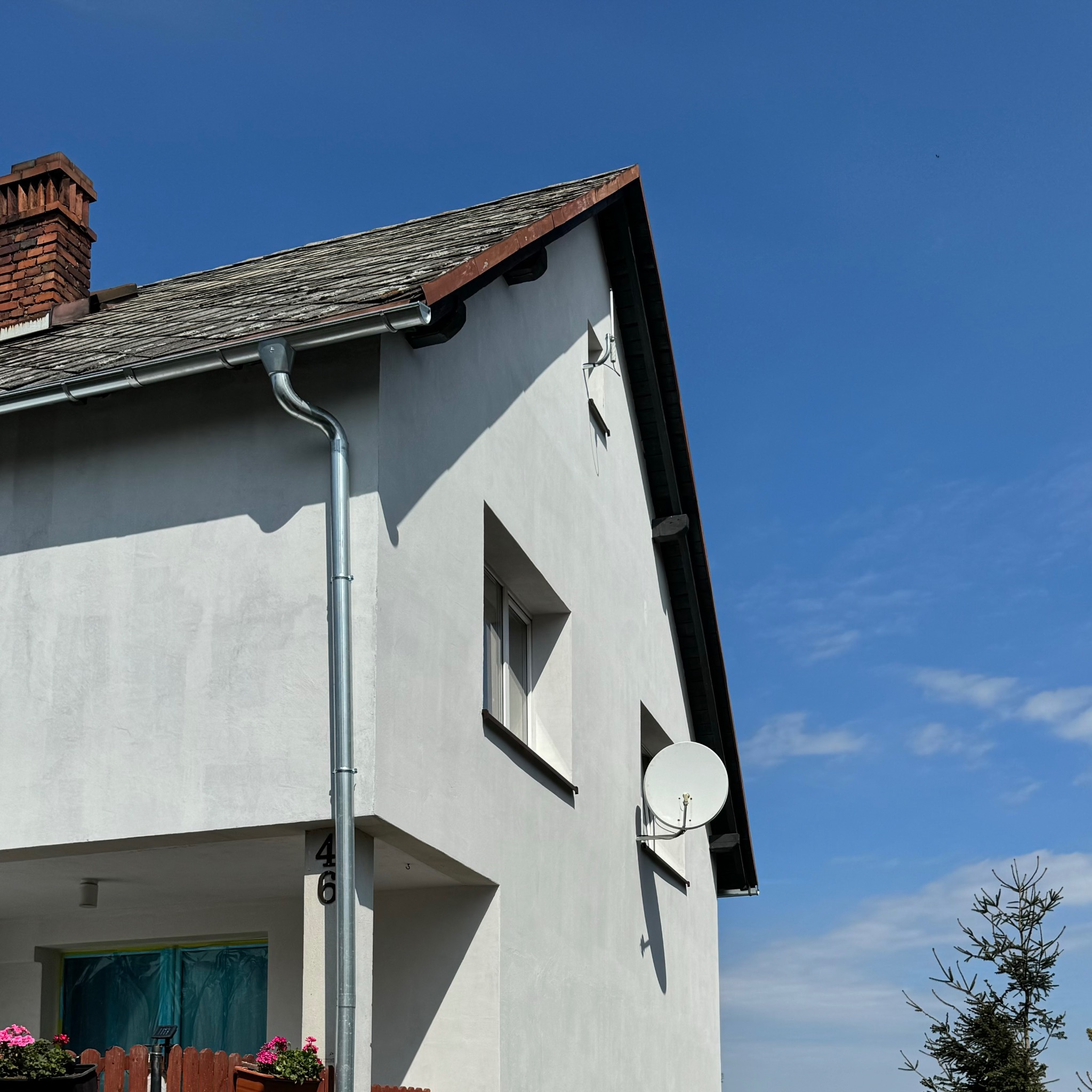 Dwupiętrowy dom z białą elewacją i szarym dachem, widoczne okna, rynna, antena satelitarna oraz komin z cegły, na tle błękitnego nieba.