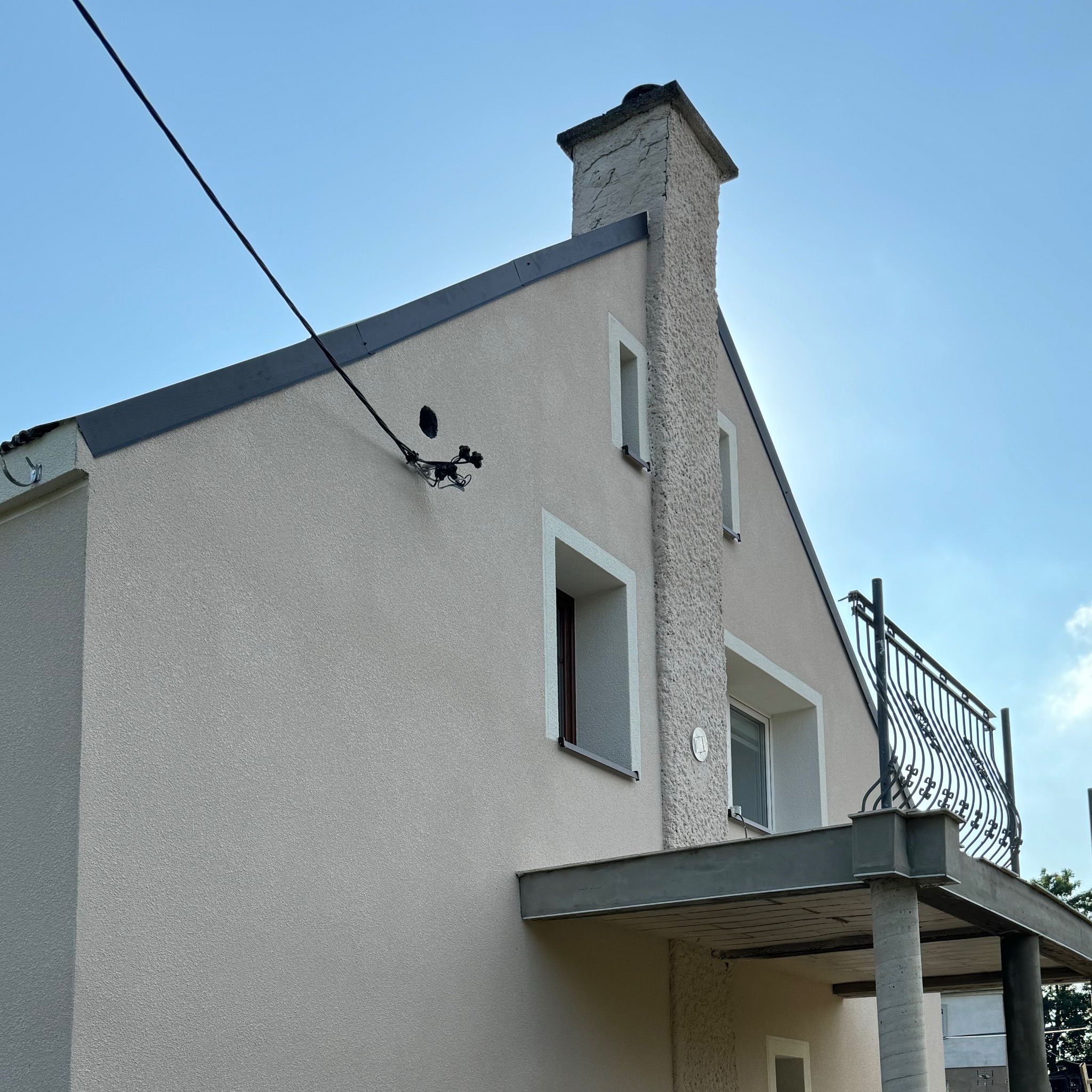 Jasna elewacja domu jednorodzinnego z dekoracyjnym kominem, balkonem z ozdobną balustradą i widocznymi przewodami elektrycznymi.