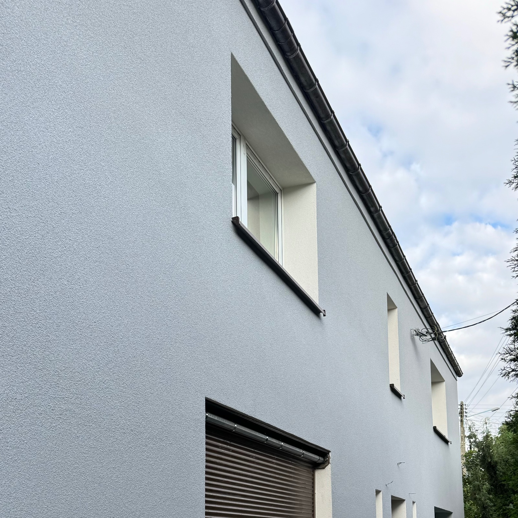 Świeżo otynkowana i pomalowana na szaro elewacja dwukondygnacyjnego budynku z widocznymi oknami, roletami zewnętrznymi i rynnami.