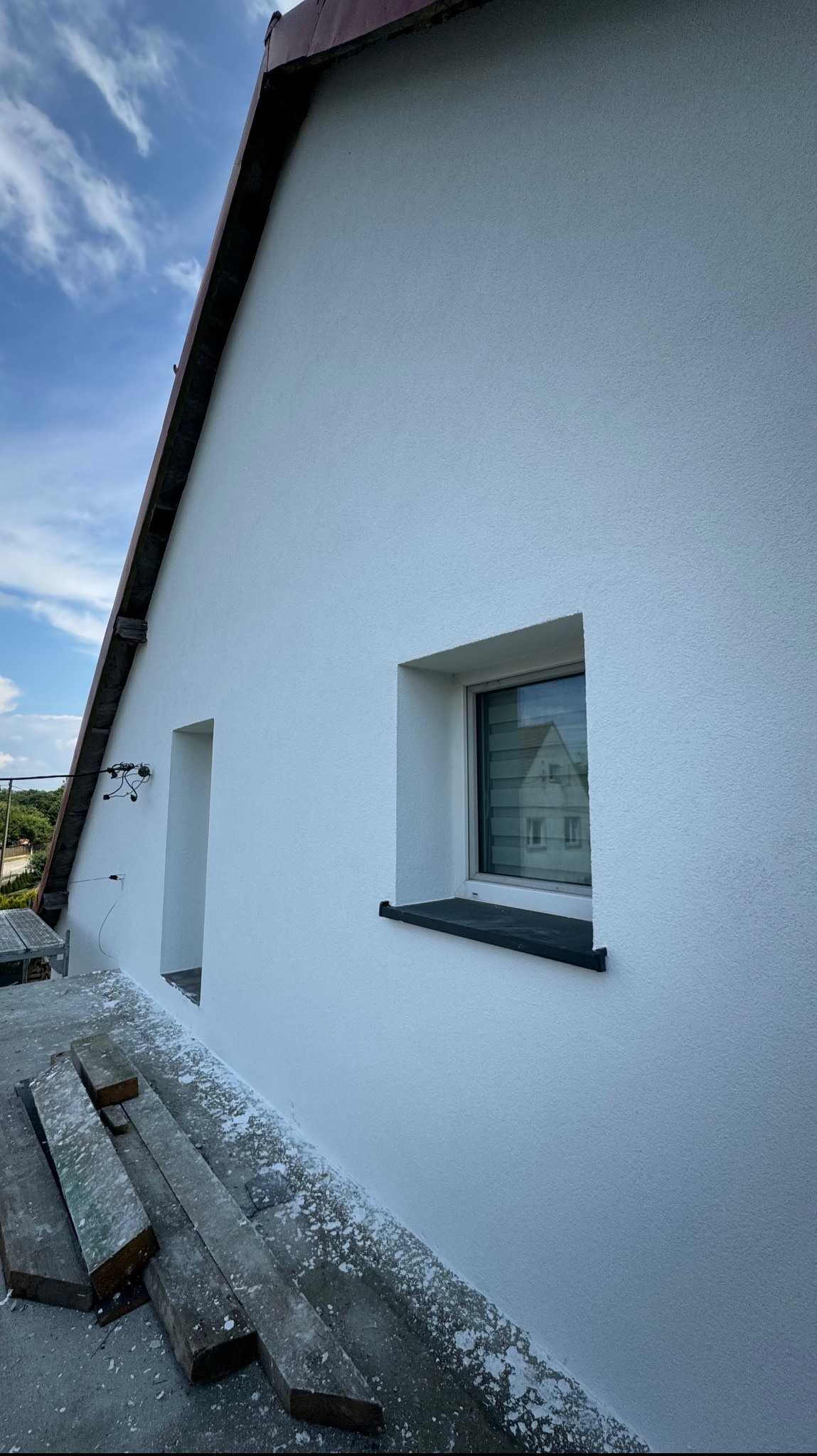 Świeżo otynkowana, biała ściana domu jednorodzinnego z widocznym oknem i drzwiami, z drewnianymi belkami leżącymi na ziemi, częściowo zabrudzonymi tynkiem, na tle błękitnego nieba.
