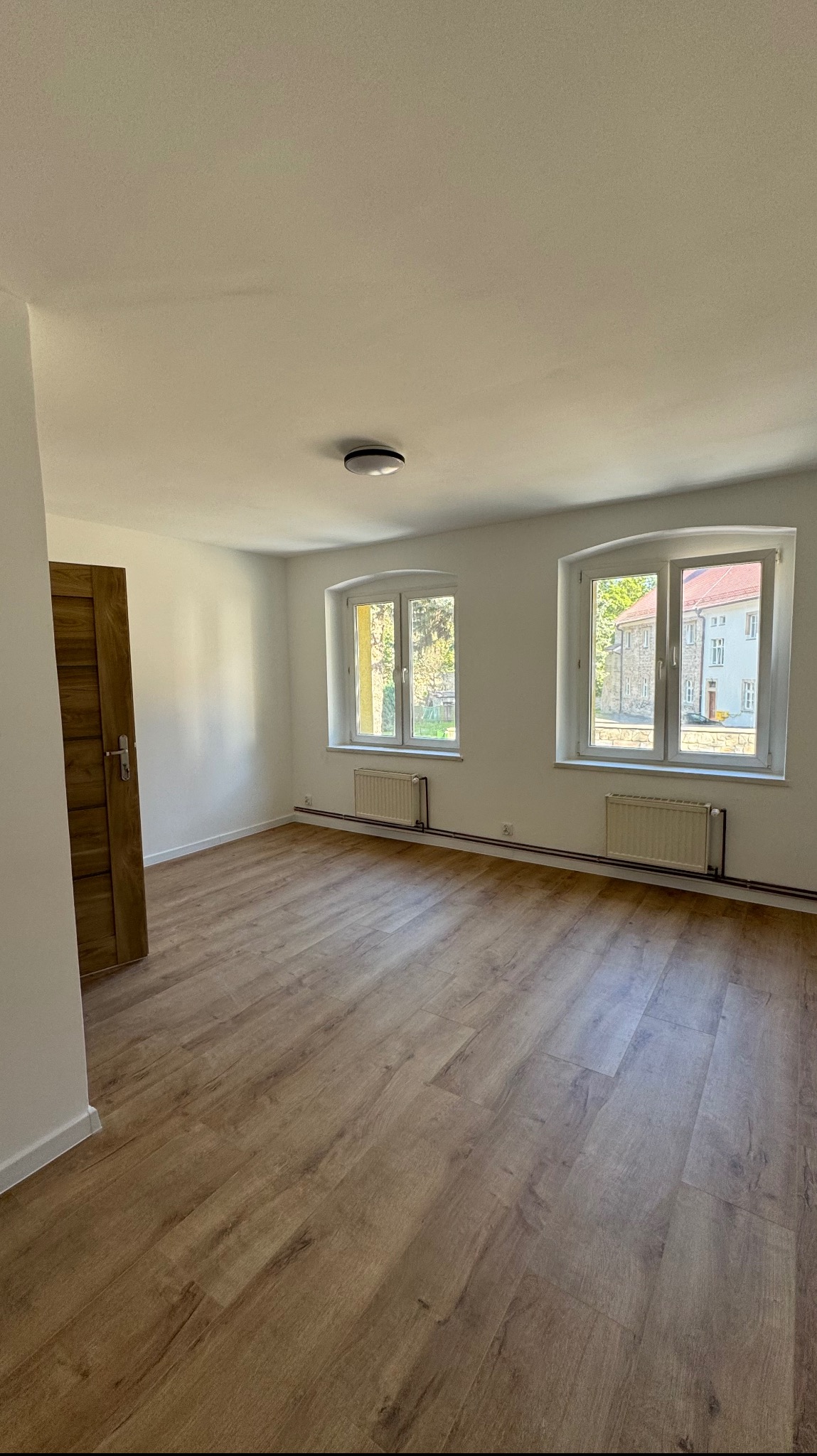 Pusty pokój po remoncie z drewnianą podłogą, dwoma oknami z widokiem na budynek i grzejnikami pod nimi, oraz drewnianymi drzwiami po lewej stronie.