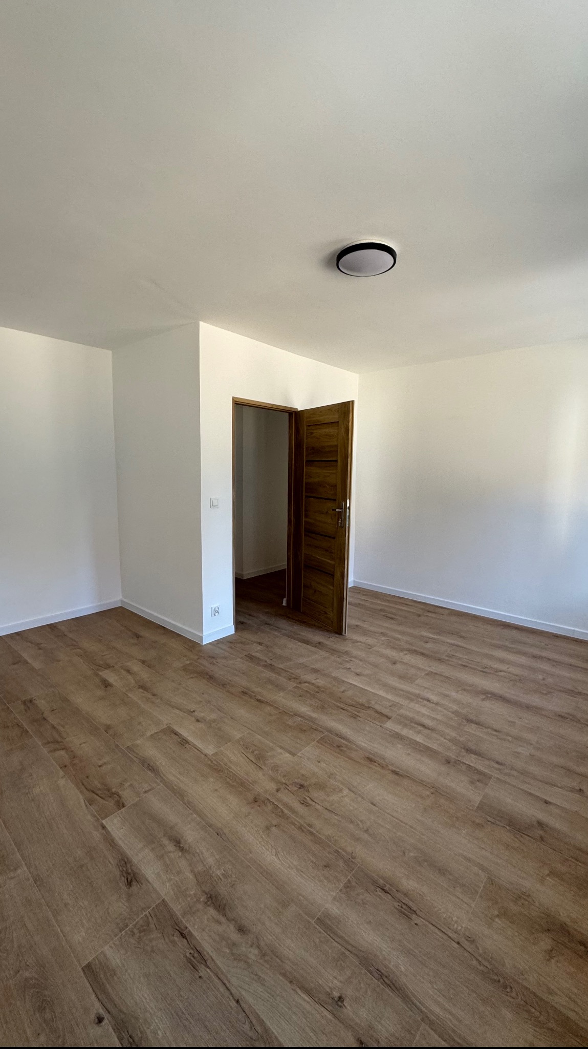 Wnętrze nowo wykończonego pokoju z panelami podłogowymi imitującymi drewno, świeżo pomalowanymi na biało ścianami i otwartymi drewnianymi drzwiami wewnętrznymi.