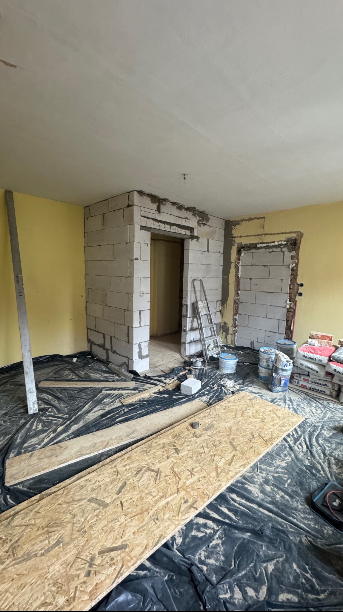 Wnętrze w trakcie prac budowlanych: wznoszenie ścianek działowych z bloczków, widoczne worki z zaprawą, płyty OSB na podłodze zabezpieczonej folią, drabina aluminiowa i narzędzia.