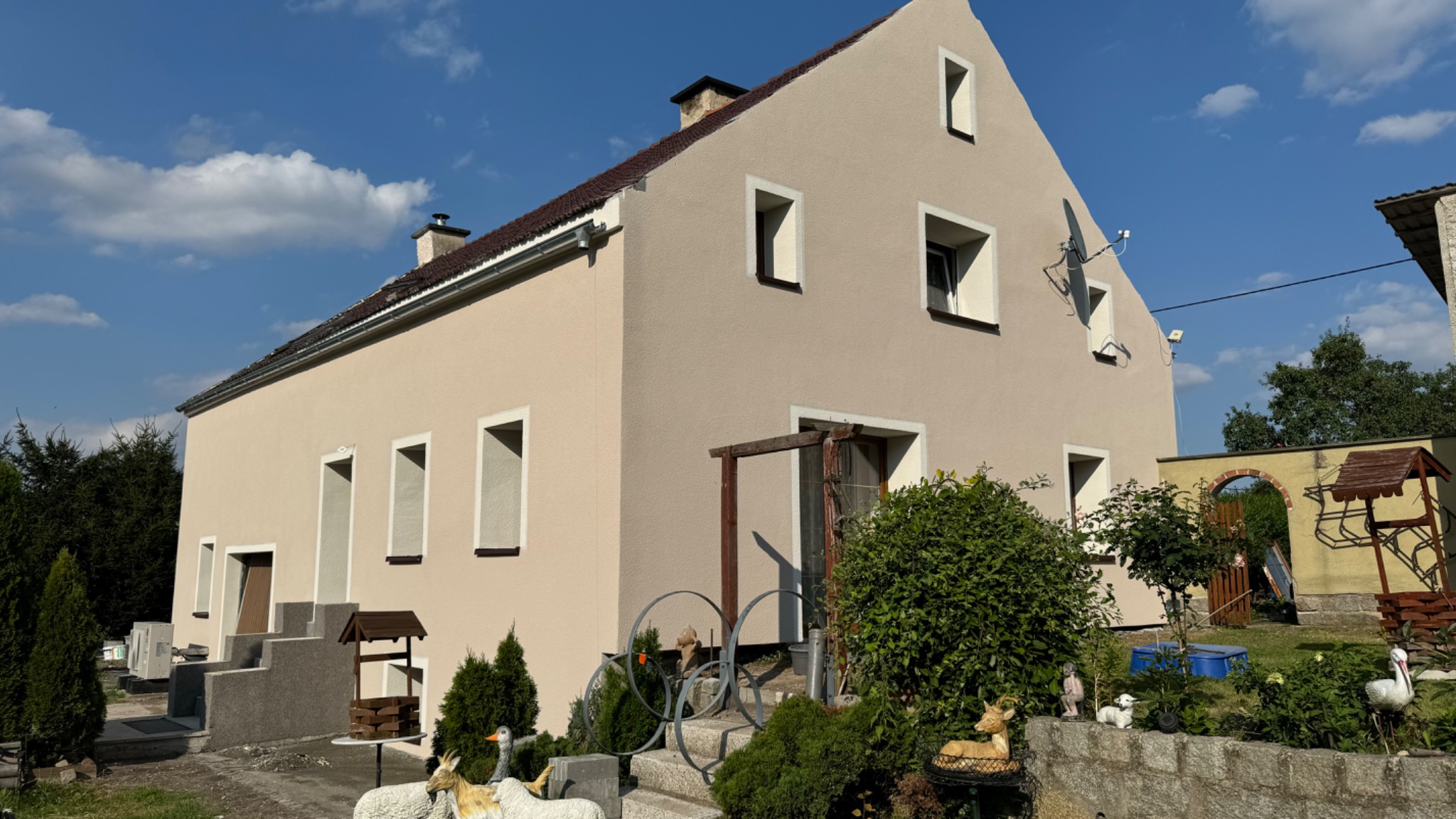 Widok z zewnątrz na dom jednorodzinny po renowacji elewacji, z dekoracyjnymi elementami ogrodowymi i satelitarną anteną na ścianie szczytowej.