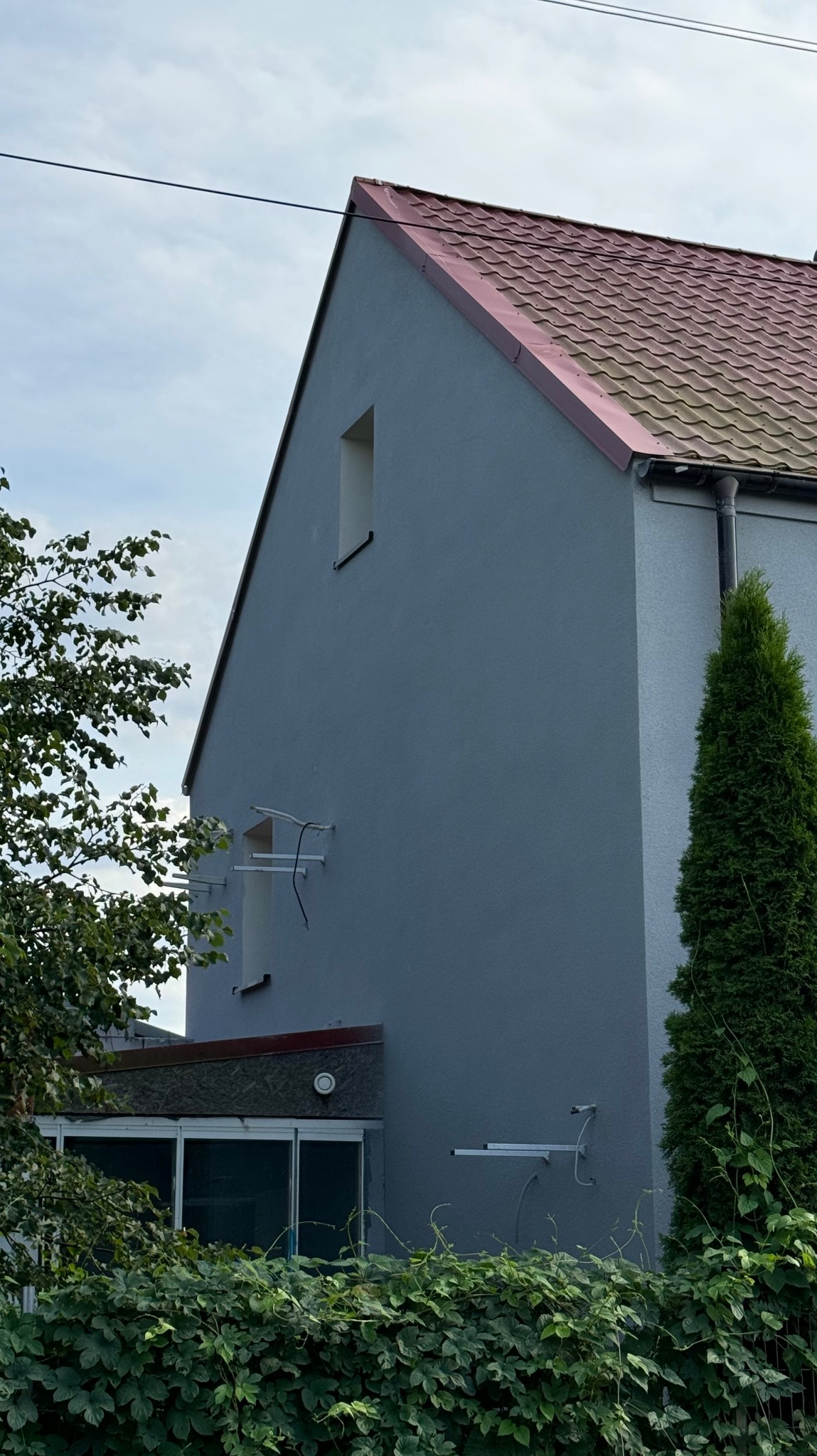 Fragment domu jednorodzinnego z szarą elewacją i czerwoną dachówką, widoczne okno na poddaszu, krzewy przed budynkiem i drzewo iglaste obok ściany, uchwyty antenowe bez anteny na ścianie