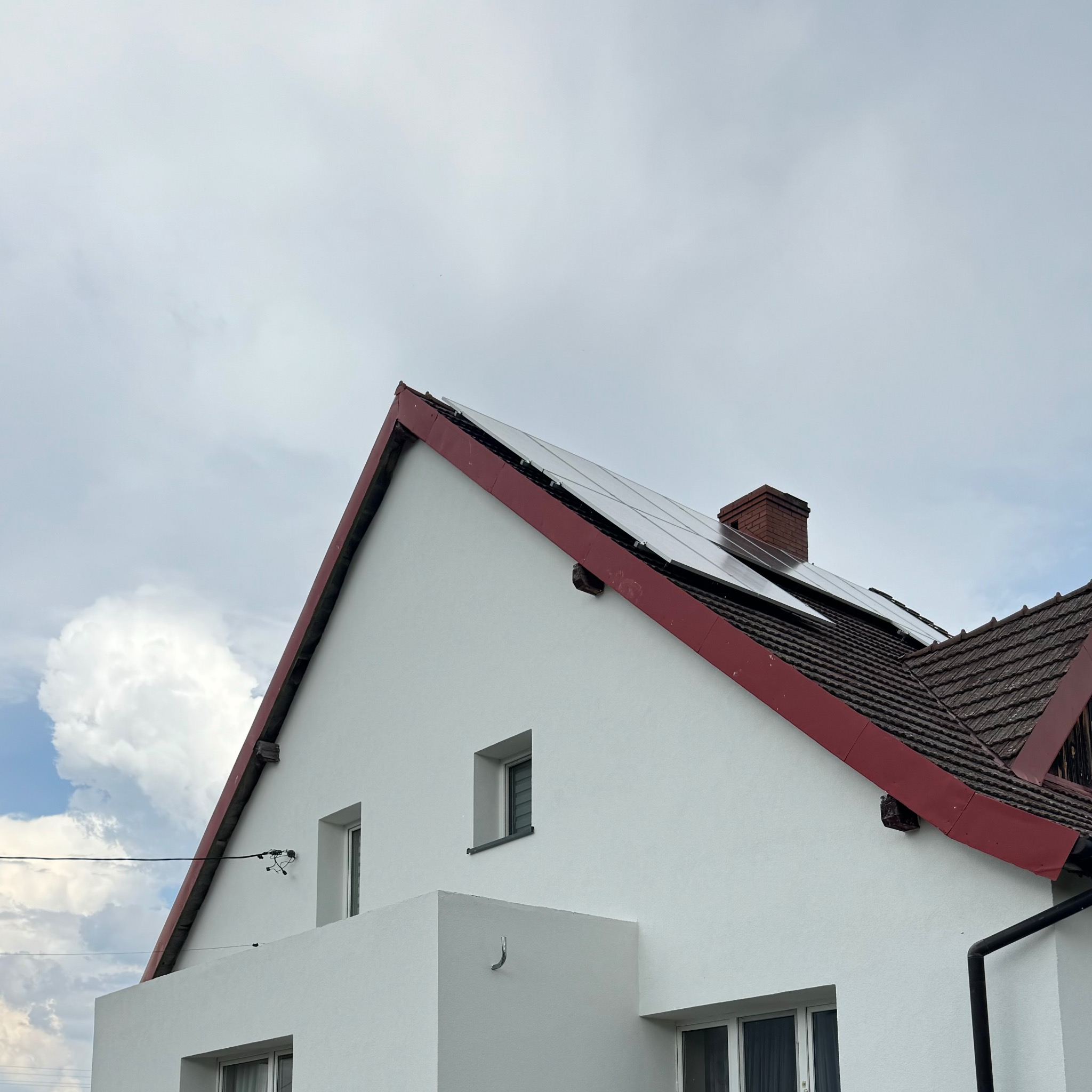 Biały dom z czerwonym dachem, panelami słonecznymi i ceglanym kominem na tle pochmurnego nieba.