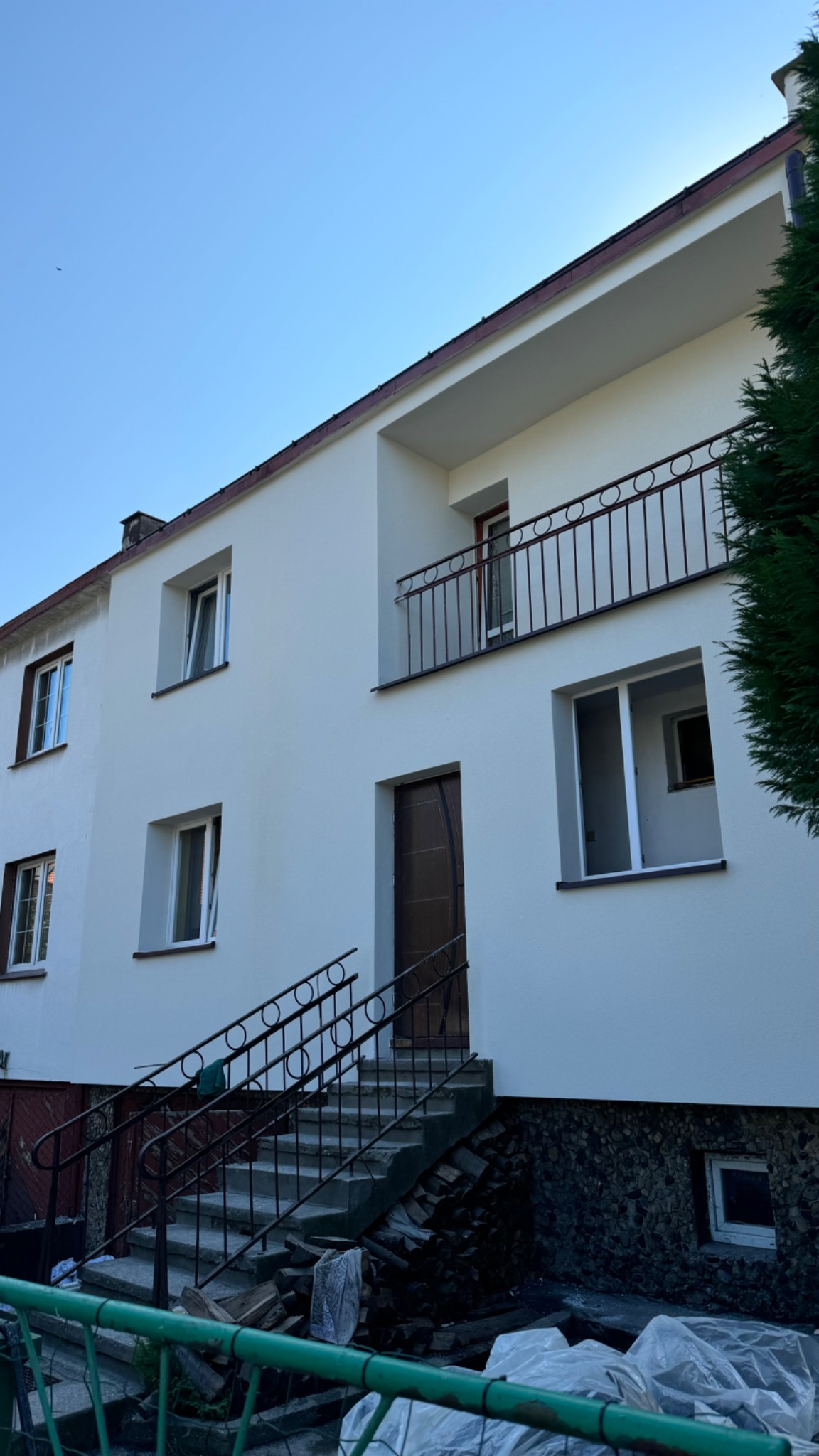 Dwupiętrowy dom z białymi ścianami po remoncie, z balkonem i schodami wejściowymi, widok z dołu na elewację, częściowo zakryty przez zielone ogrodzenie i drewno opałowe.