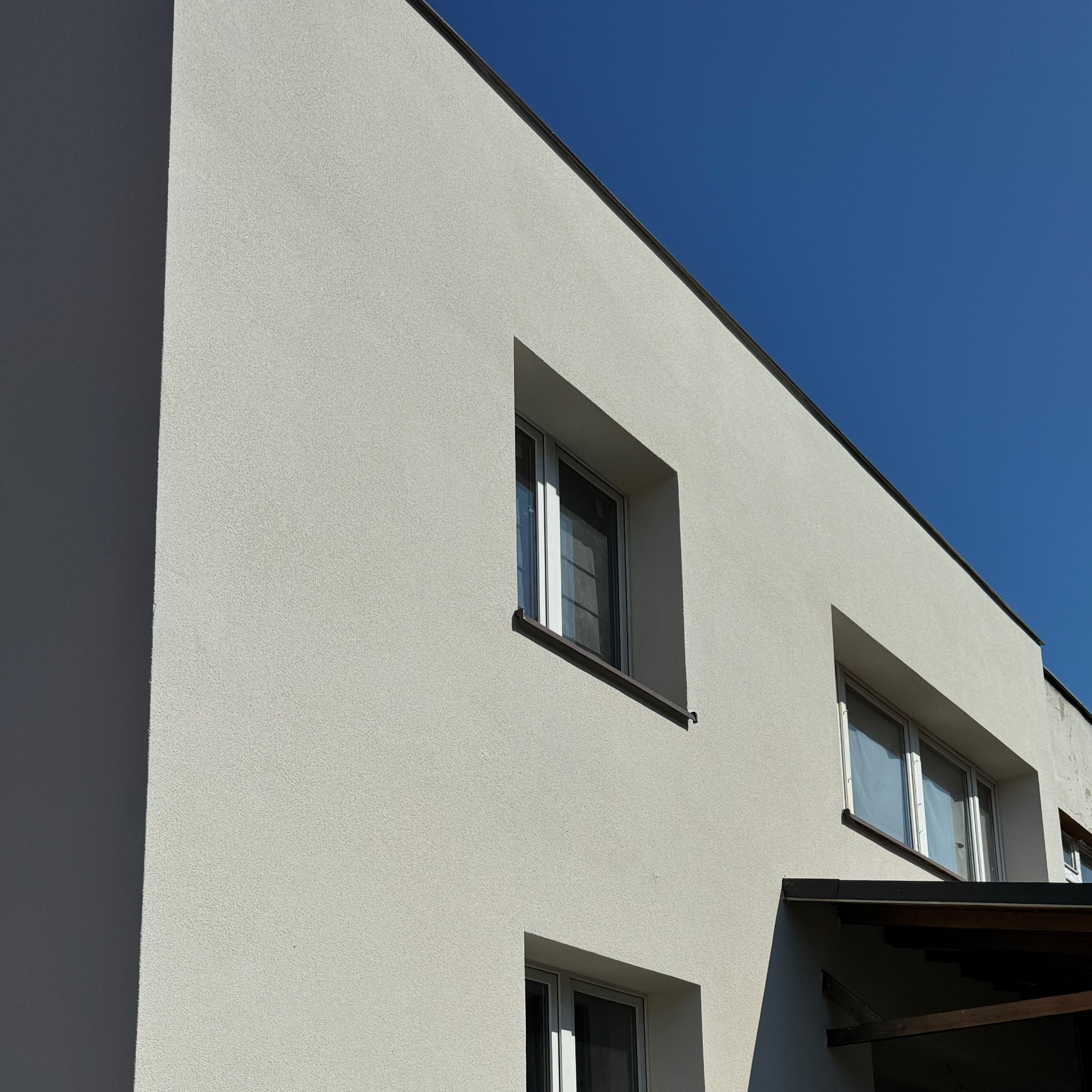 Fragment nowoczesnego domu z jasną, strukturalną elewacją i oknami z ciemnymi ramami, widziany pod kątem na tle błękitnego nieba.