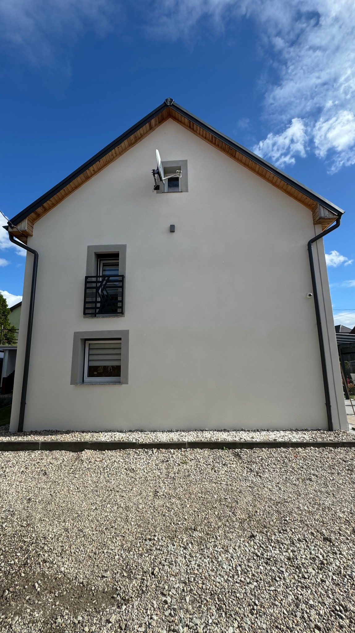 Dwukondygnacyjny dom jednorodzinny z jasną elewacją, widoczne okna z czarną balustradą i roletami, antena satelitarna na poddaszu, rynny w kolorze grafitowym, podłoże wysypane drobnym kamieniem...