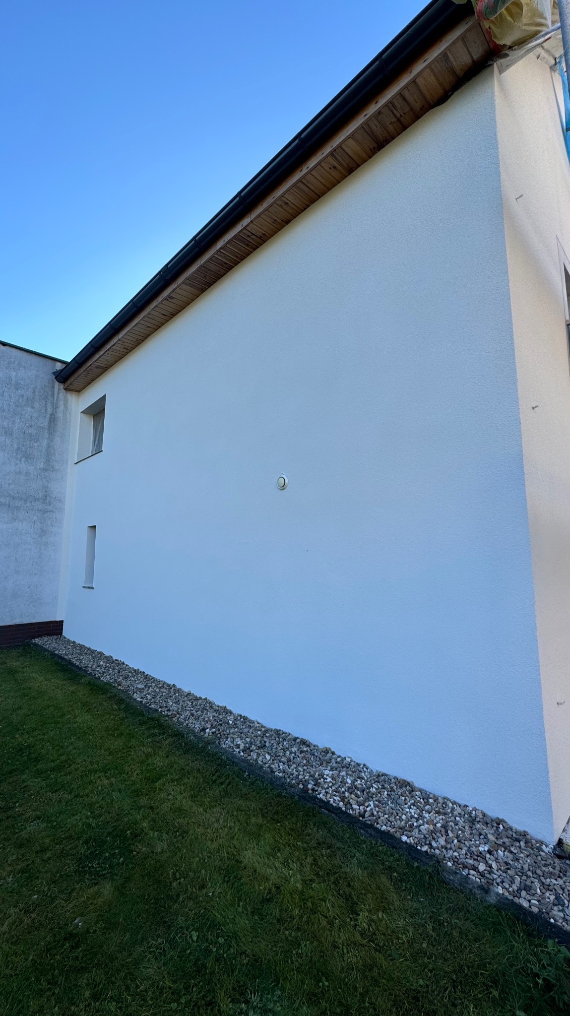 Świeżo otynkowana i pomalowana na biało ściana domu jednorodzinnego z widocznym fragmentem elewacji sąsiedniego budynku, trawnikiem i kamiennym obrzeżem.