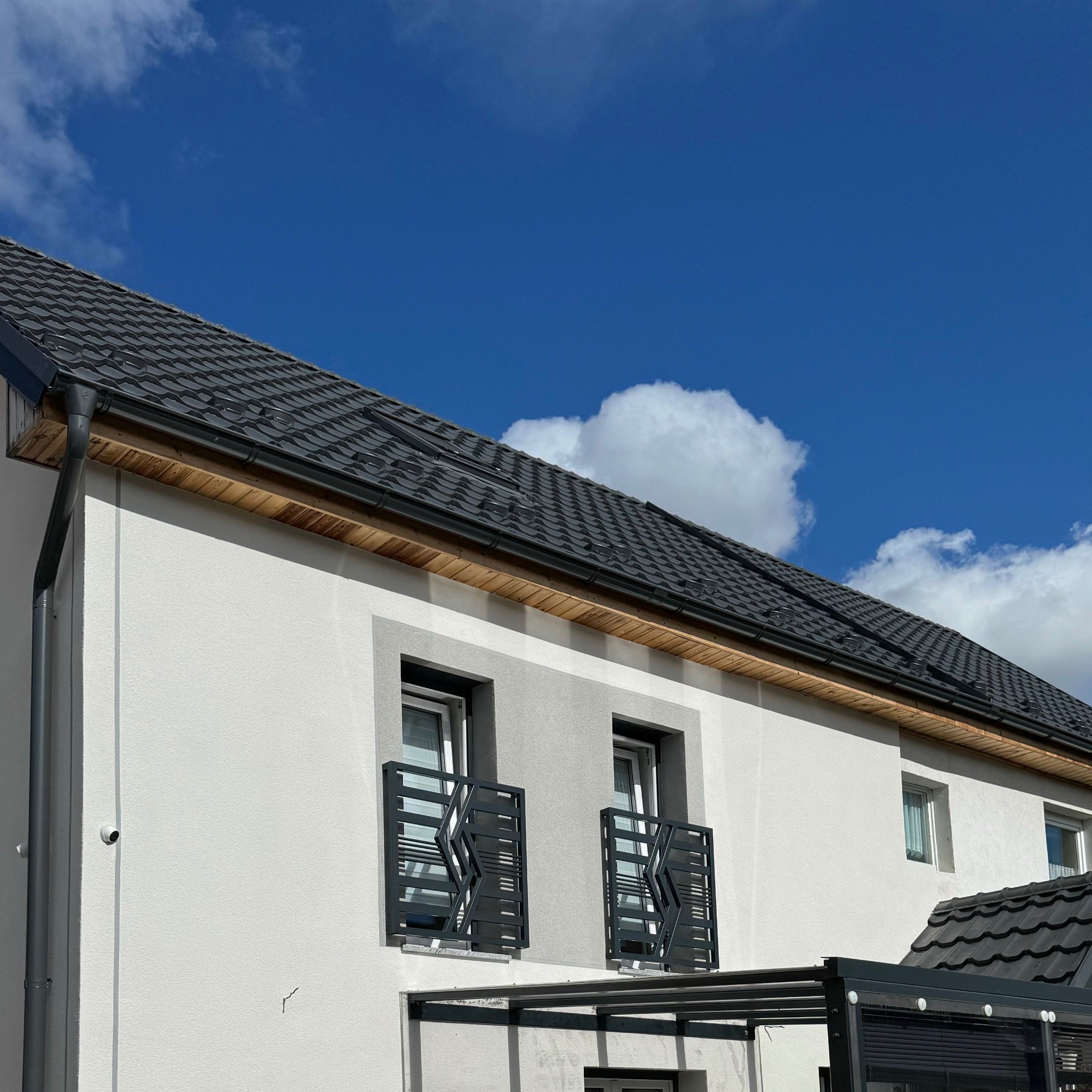 Nowoczesny dom jednorodzinny z jasną elewacją, czarną dachówką i balkonami z dekoracyjnymi balustradami na tle błękitnego nieba z chmurami.