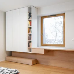 Nowoczesny pokój z białą szafą w zabudowie, drewnianym biurkiem pod oknem z widokiem na zimowy krajobraz i minimalistyczną lampką, uzupełniony o drewnianą podłogę i fragment wzorzystej narzuty.