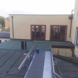 Zielony dach z widocznymi śladami po naprawach, metalowy pomost z barierkami, butle gazowe i kominy wentylacyjne na tle budynku z oknami.