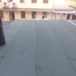 Widok na płaski dach pokryty papą termozgrzewalną w kolorze zielonym, z widocznymi rolkami papy marki Lemar w lewym rogu i budynkami w tle.