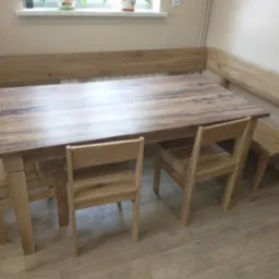 Drewniany stół z krzesłami i narożną ławą w jasnym odcieniu, widoczne słoje drewna na blacie stołu.