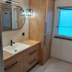 Nowoczesna łazienka z drewnopodobną zabudową szafek pod umywalką, wysoką szafą i okrągłym lustrem z podświetleniem LED, uzupełniona wiszącą lampą z czarną ramą.