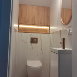 Nowoczesna łazienka po remoncie: wisząca toaleta, szafka z umywalką, lustro w złotej ramie, drewniane szafki podświetlone LED, marmurowe płytki z żyłami w kolorze złota.