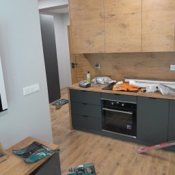 IDEAL flizowanie&łazienki - Montaż kuchni w trakcie: widoczne szafki górne z drewnopodobnym fornirem, szare szafki dolne z piekarnikiem, narzędzia i materiały budowlane na blacie oraz na podłodze z paneli.