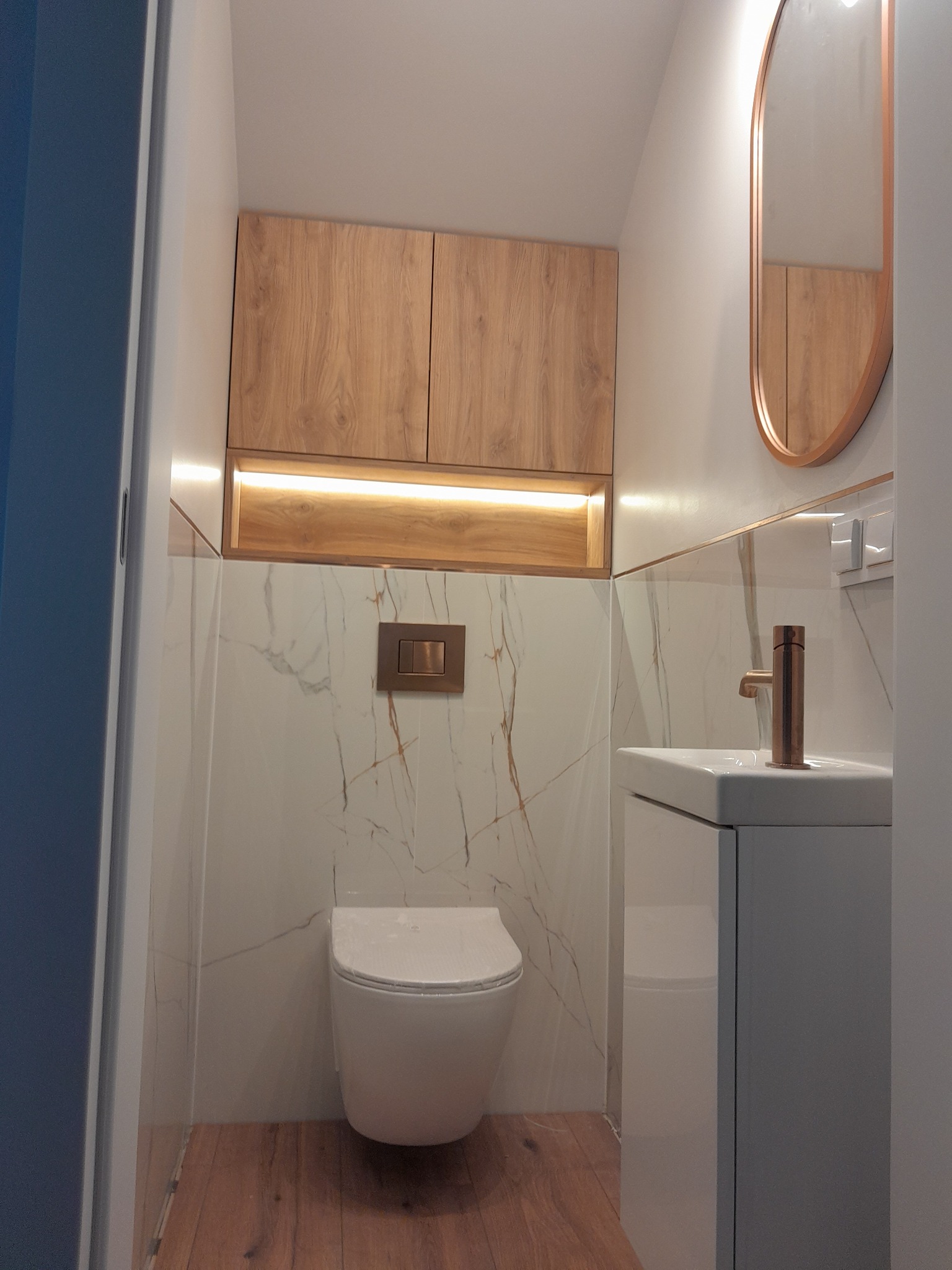Nowoczesna łazienka po remoncie: wisząca toaleta, szafka z umywalką, lustro w złotej ramie, drewniane szafki podświetlone LED, marmurowe płytki z żyłami w kolorze złota.