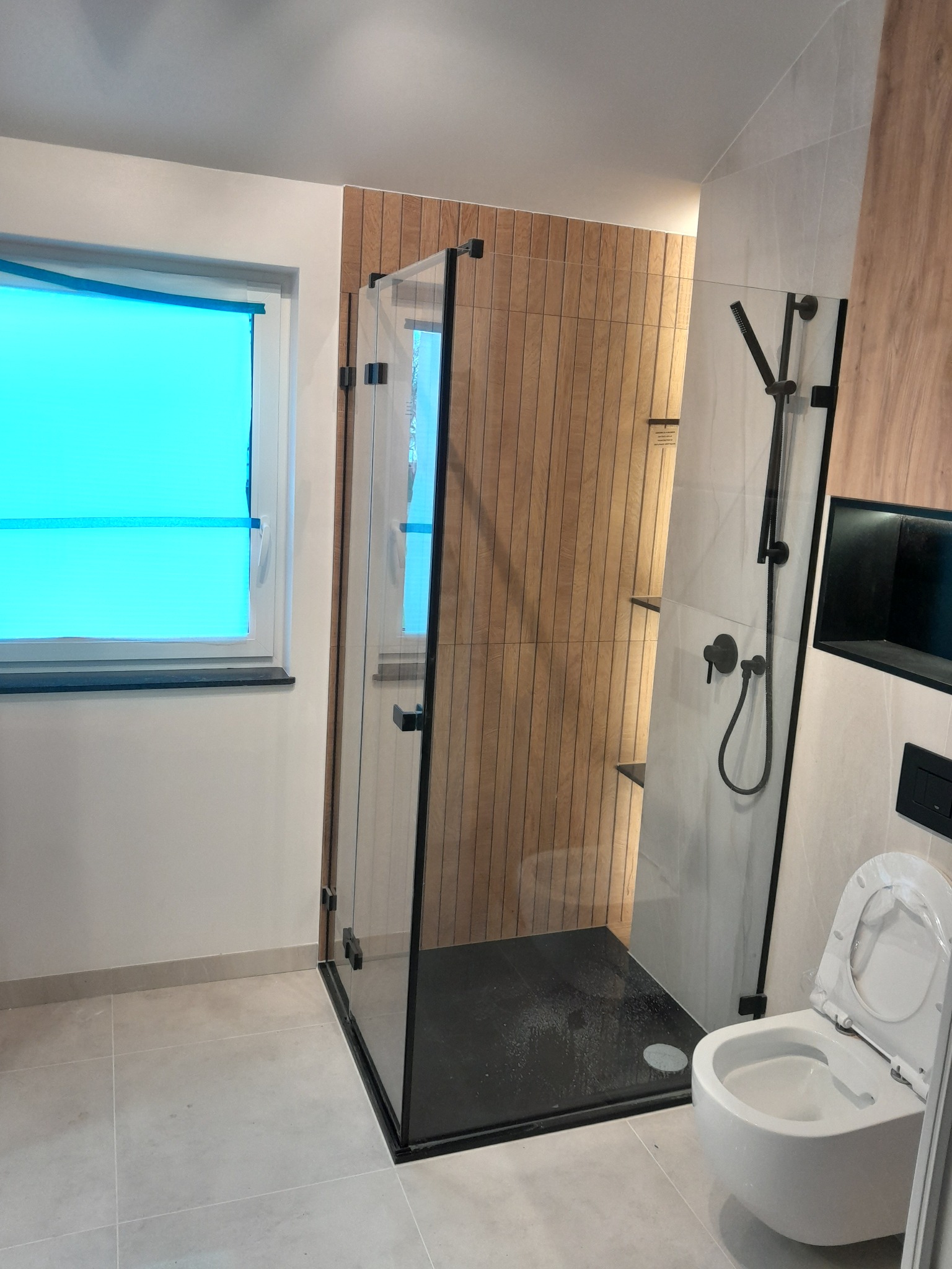 Nowoczesna łazienka z kabiną prysznicową typu walk-in z czarnymi profilami, drewnopodobną ścianą i wiszącą miską WC, okno z niebieską folią ochronną.
