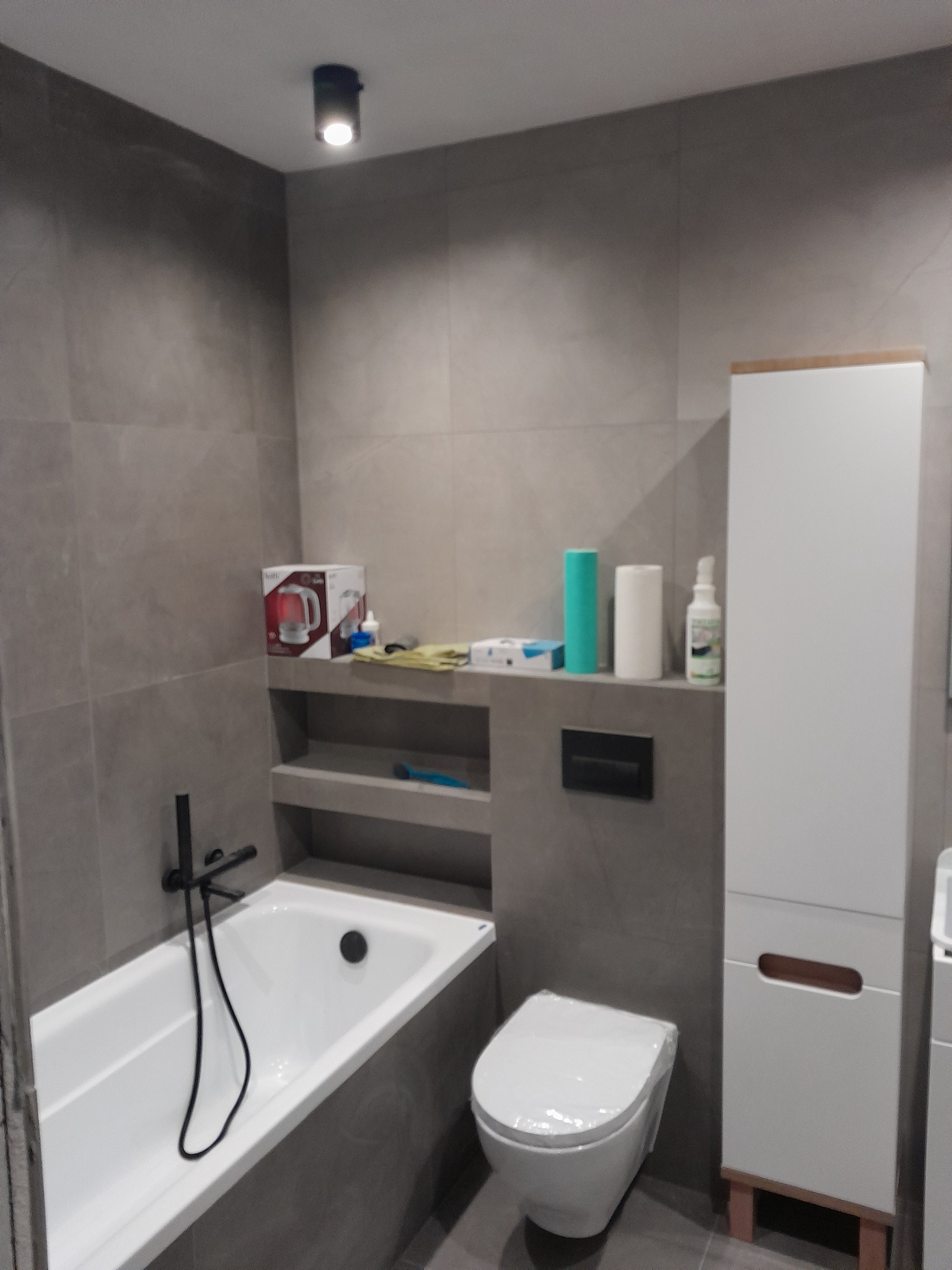 Nowoczesna łazienka w szarościach z białą wanną, wiszącą toaletą w folii ochronnej i szafką, oświetlona punktowo, z akcentami przedmiotów na półce.