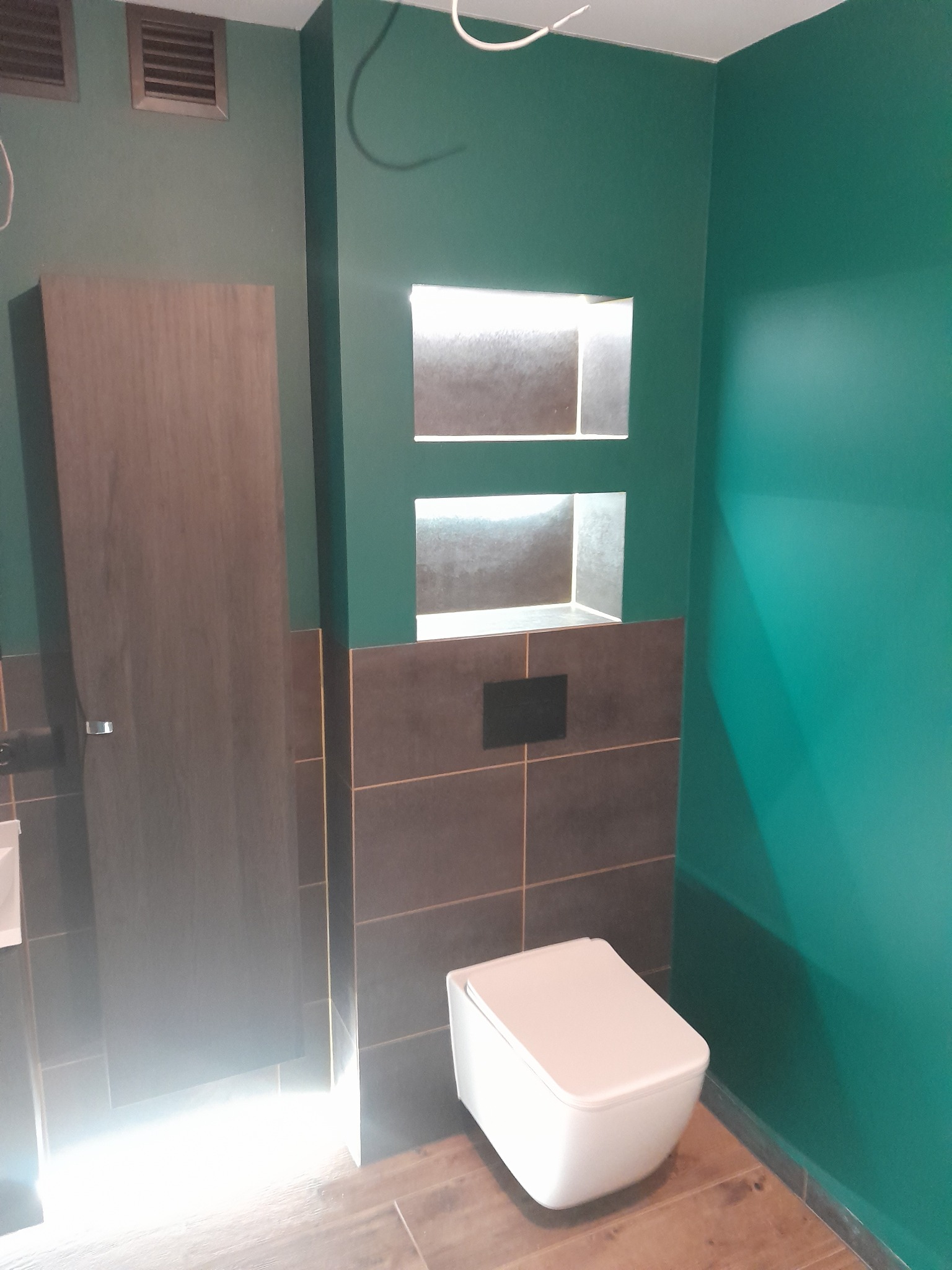 Nowoczesna łazienka z zielonymi ścianami, zabudowaną toaletą, brązowymi płytkami i wnękami z podświetleniem LED.