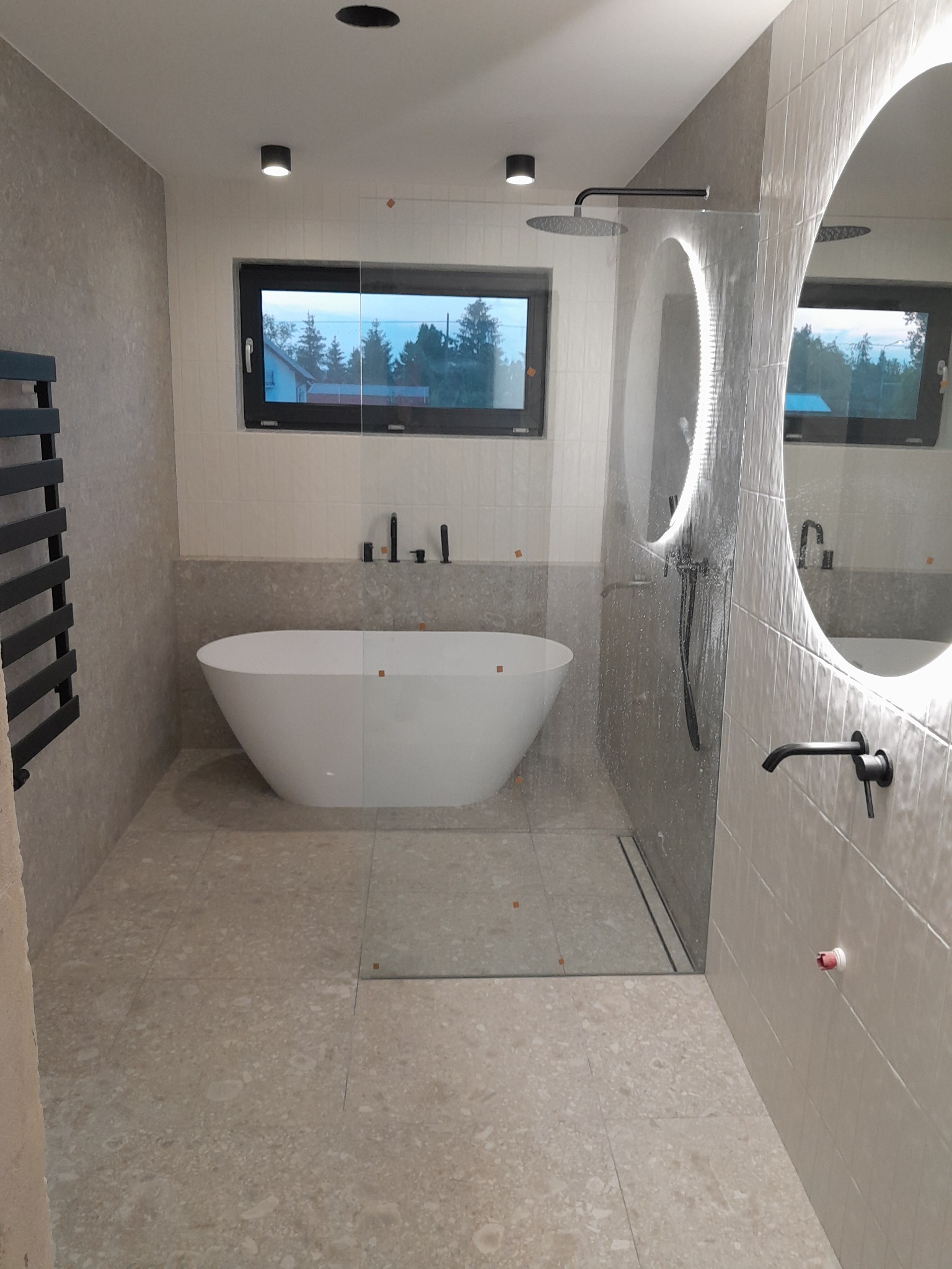 Nowoczesna łazienka z wolnostojącą białą wanną, oświetlonym lustrem, czarną armaturą i dużym oknem z widokiem na zieleń.