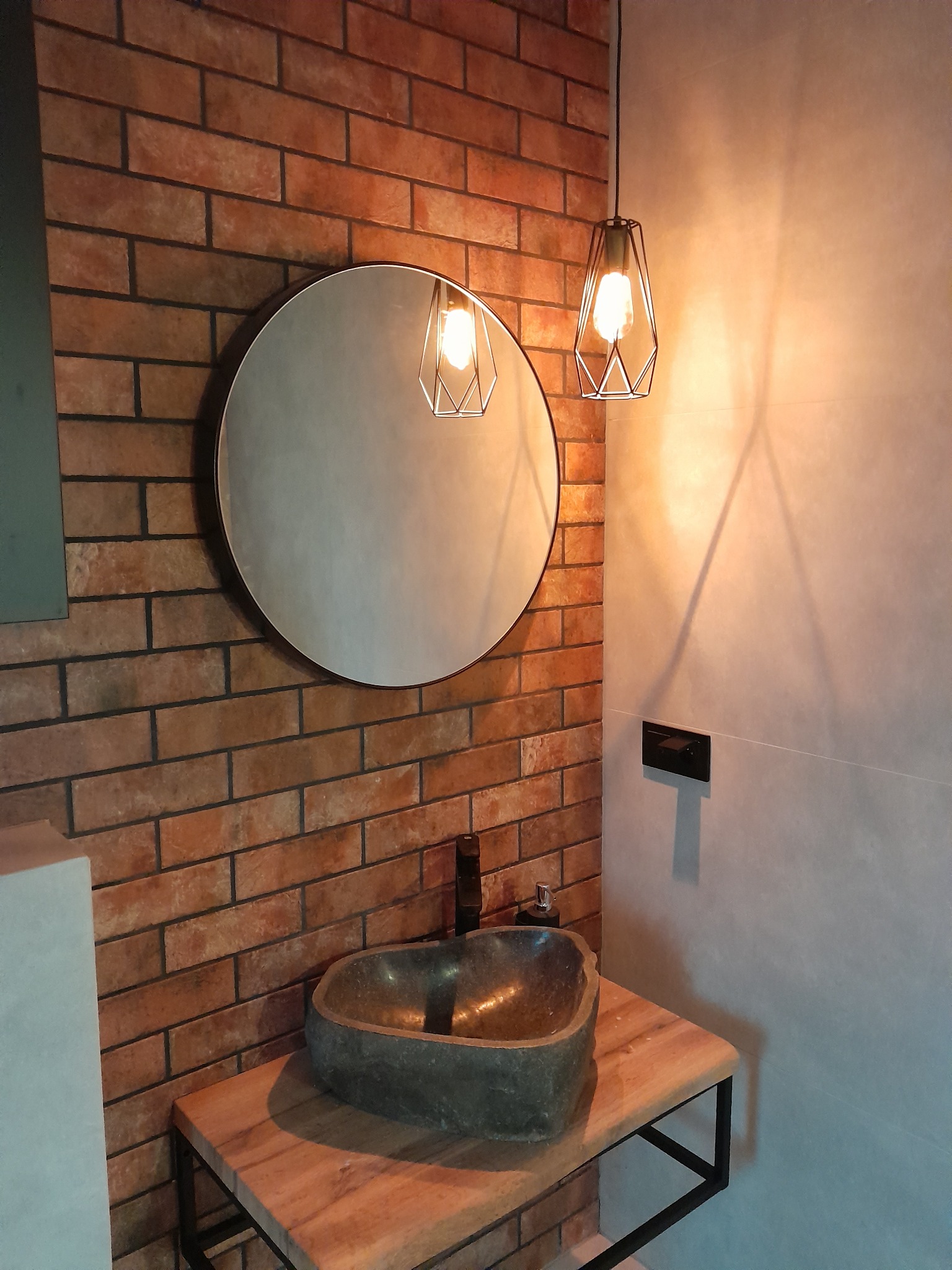 Nowoczesna łazienka z umywalką z naturalnego kamienia, okrągłym lustrem i oświetleniem w stylu industrialnym na tle ściany z cegły.