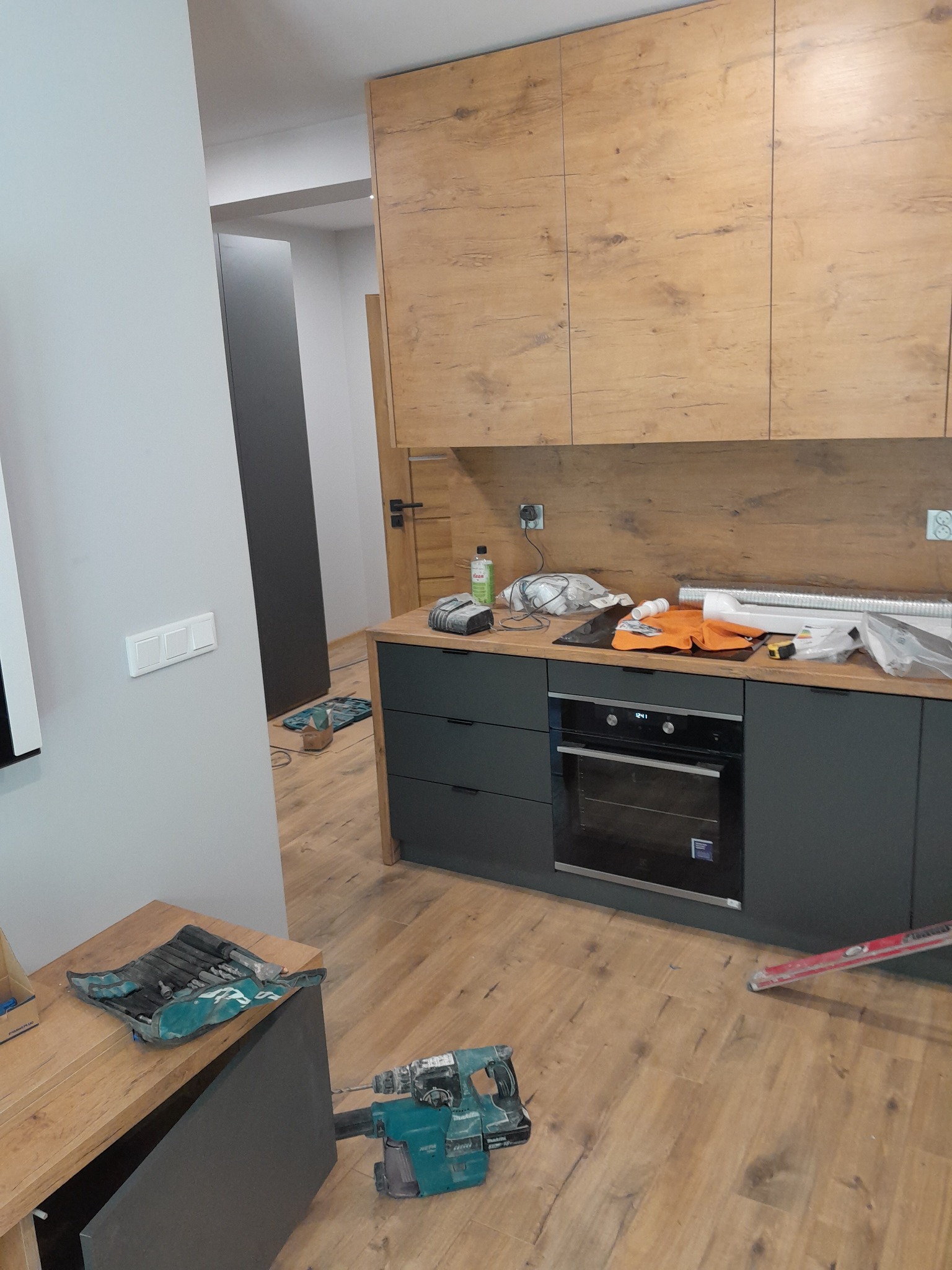 Montaż kuchni w trakcie: widoczne szafki górne z drewnopodobnym fornirem, szare szafki dolne z piekarnikiem, narzędzia i materiały budowlane na blacie oraz na podłodze z paneli.