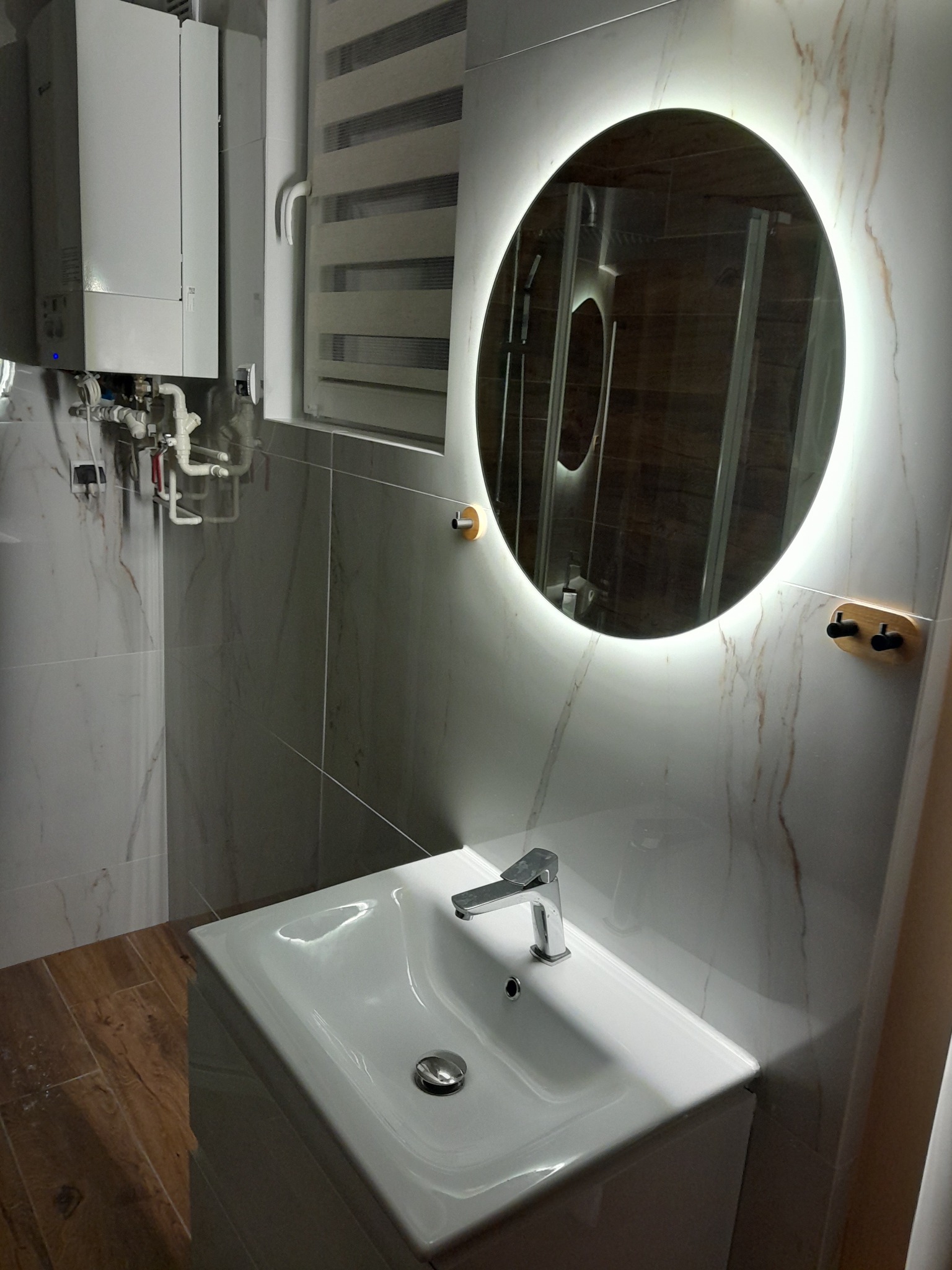 Nowoczesna łazienka z umywalką nablatową, okrągłym lustrem z podświetleniem LED, marmurowymi płytkami na ścianach i drewnopodobną podłogą, widoczny bojler oraz roleta okienna.