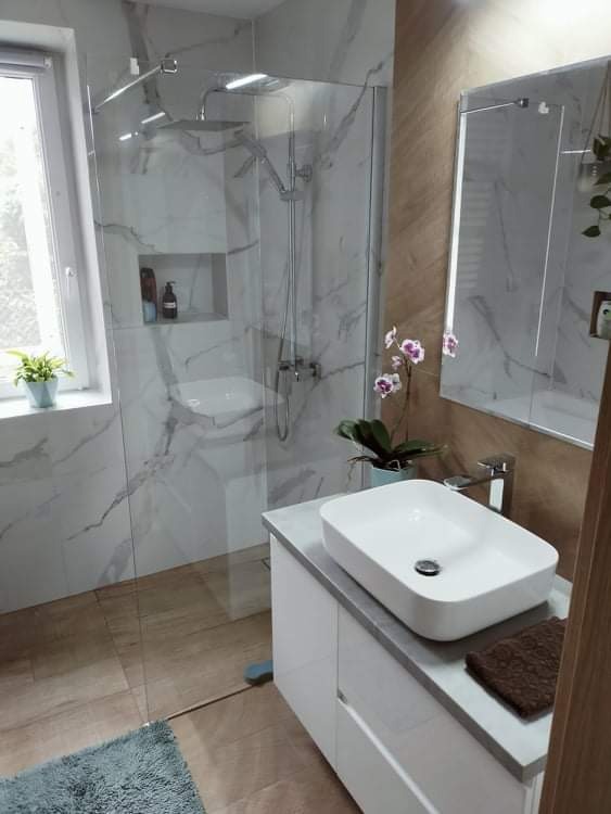 Nowoczesna łazienka z kabiną prysznicową typu walk-in, marmurowymi płytkami na ścianach, drewnopodobną podłogą, białą szafką z umywalką nablatową, lustrem i storczykiem.