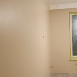 Usługi Ogólnobudowlane Sieczkarz Patryk - Świeżo pomalowane ściany na jasny beż, widoczne wystające kable ze ściany i sufitu, okno zabezpieczone folią malarską, surowa podłoga, w rogu widoczna kuweta malarska z folią.