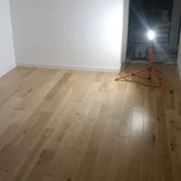 Ułożone panele podłogowe w jasnym odcieniu drewna, widoczna lampa budowlana na trójnogu, białe ściany i listwy przypodłogowe.