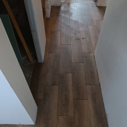 Usługi Ogólnobudowlane Sieczkarz Patryk - Ułożone brązowe panele podłogowe imitujące drewno w korytarzu podczas prac wykończeniowych, widoczne narzędzia i materiały budowlane w tle.