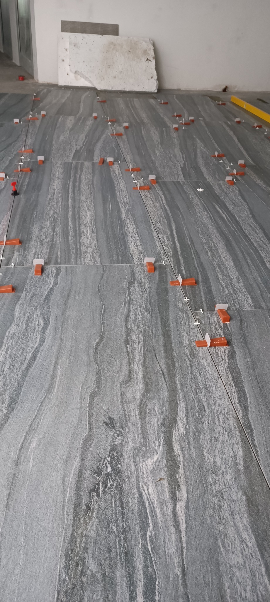 Układanie szarych płytek podłogowych imitujących kamień, z użyciem pomarańczowych klipsów poziomujących i białych krzyżyków dystansowych.