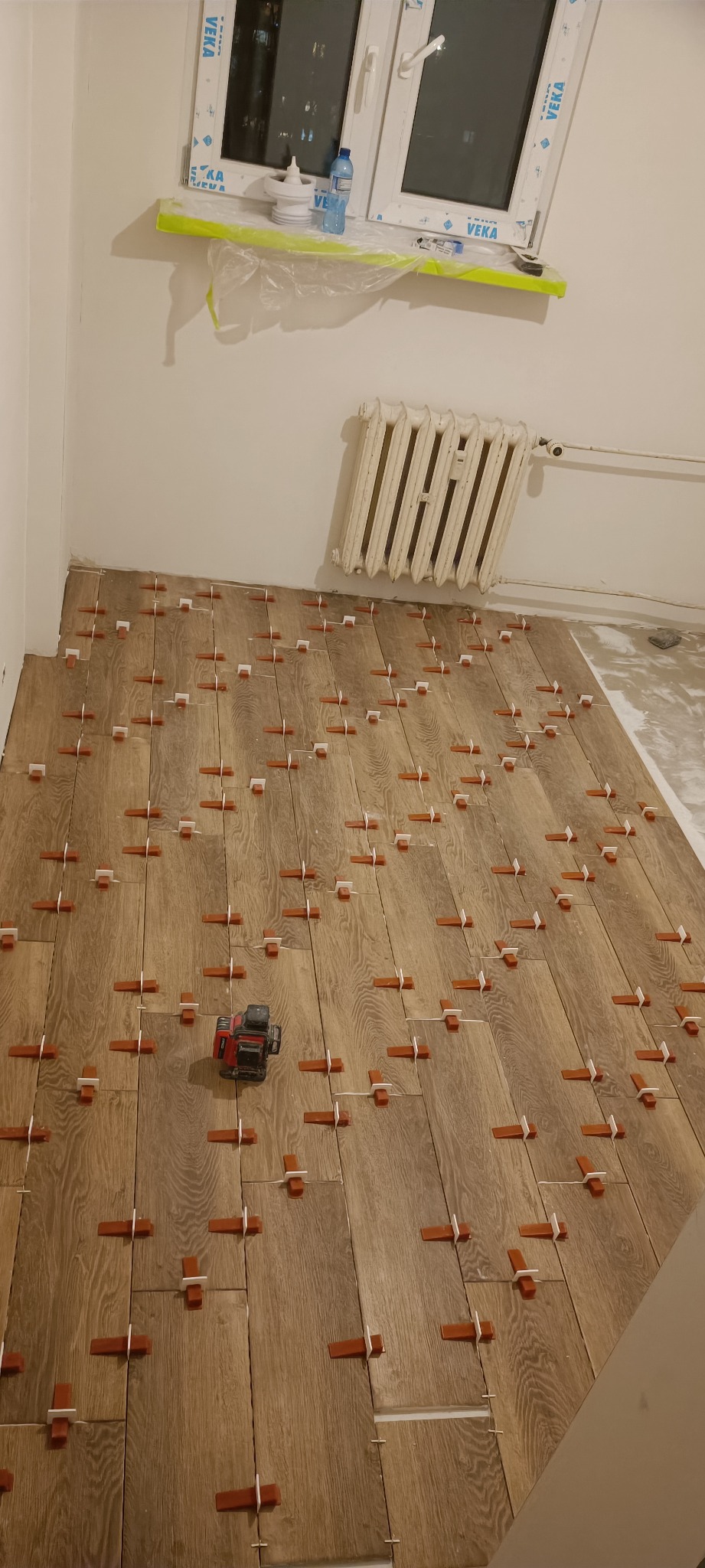 Układanie paneli podłogowych w pokoju z użyciem poziomicy laserowej i krzyżyków dystansowych, widoczne przygotowane podłoże i grzejnik ścienny.