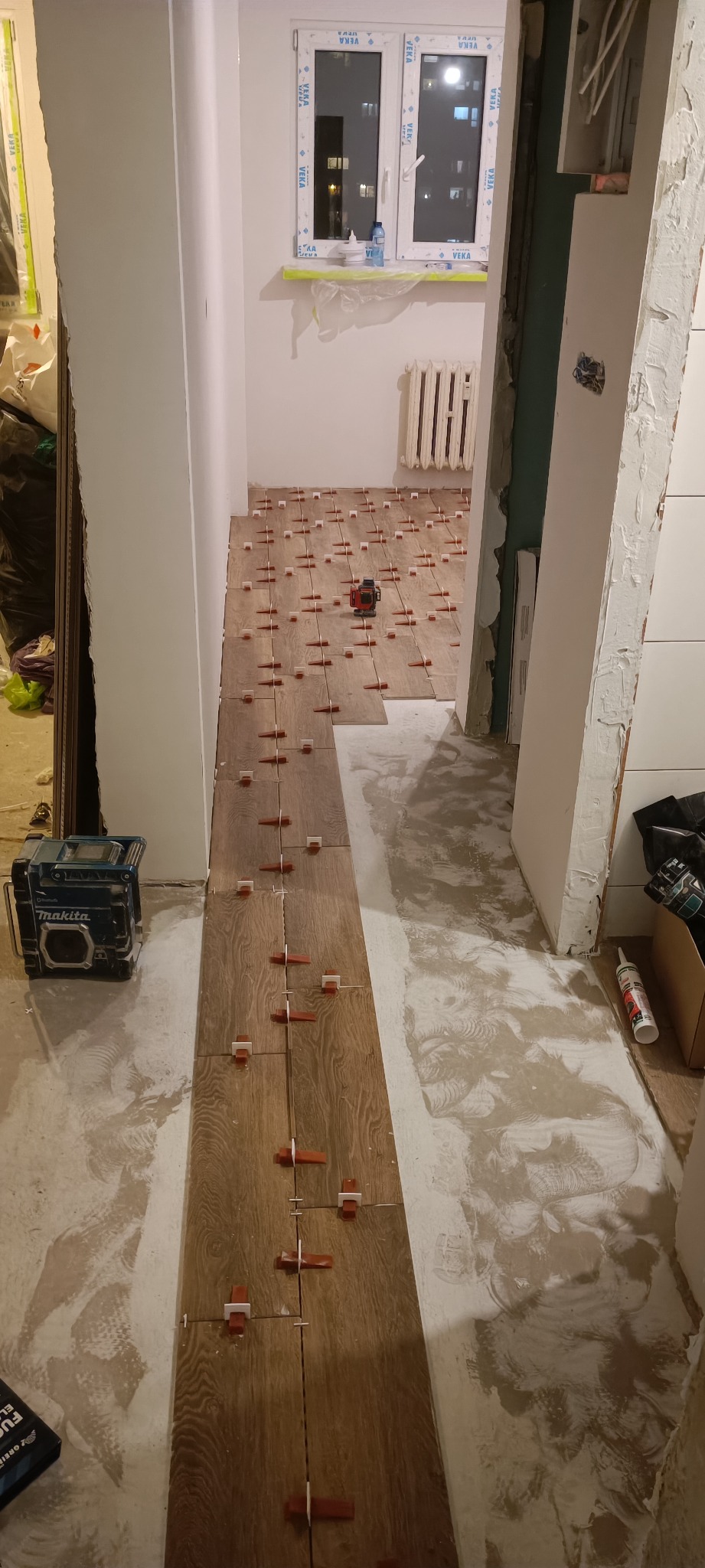 Układanie paneli podłogowych z drewnopodobnym wzorem, z użyciem poziomicy laserowej Makita i plastikowych krzyżyków dystansowych, widoczne przygotowane podłoże i grzejnik w tle.