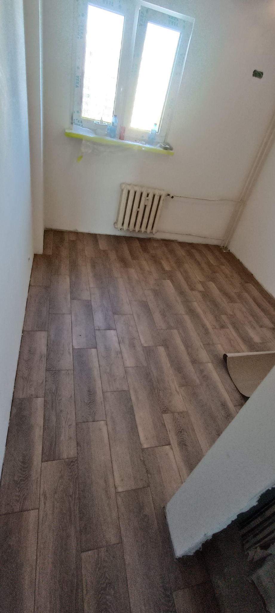 Ułożone panele podłogowe imitujące drewno w jasnym pokoju z grzejnikiem i oknem, widoczny fragment rolki podkładu.