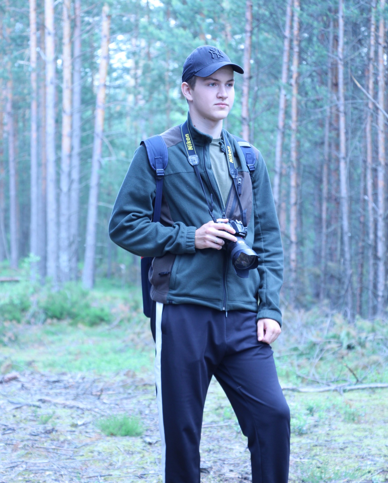 Młody mężczyzna w lesie, ubrany w polar, dresy i czapkę z daszkiem, trzyma aparat fotograficzny Nikon na szyi i patrzy w dal.