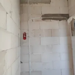 Surowa ściana z betonu komórkowego z widoczną instalacją elektryczną w postaci białych przewodów przymocowanych klamrami, czerwona puszka podwójnego gniazdka elektrycznego. Na górze ściany zwój kabli.