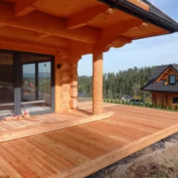 Drewniany taras w trakcie budowy przy domu z bali, widoczne deski podłogowe i materiały budowlane, w tle las i dom z ciemnym dachem.