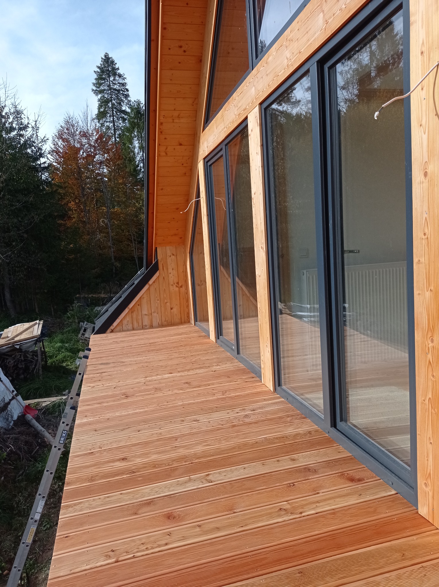 Nowy drewniany taras z desek ryflowanych przy domu z dużymi oknami przesuwnymi, widok na las jesienny, drabina oparta o taras.