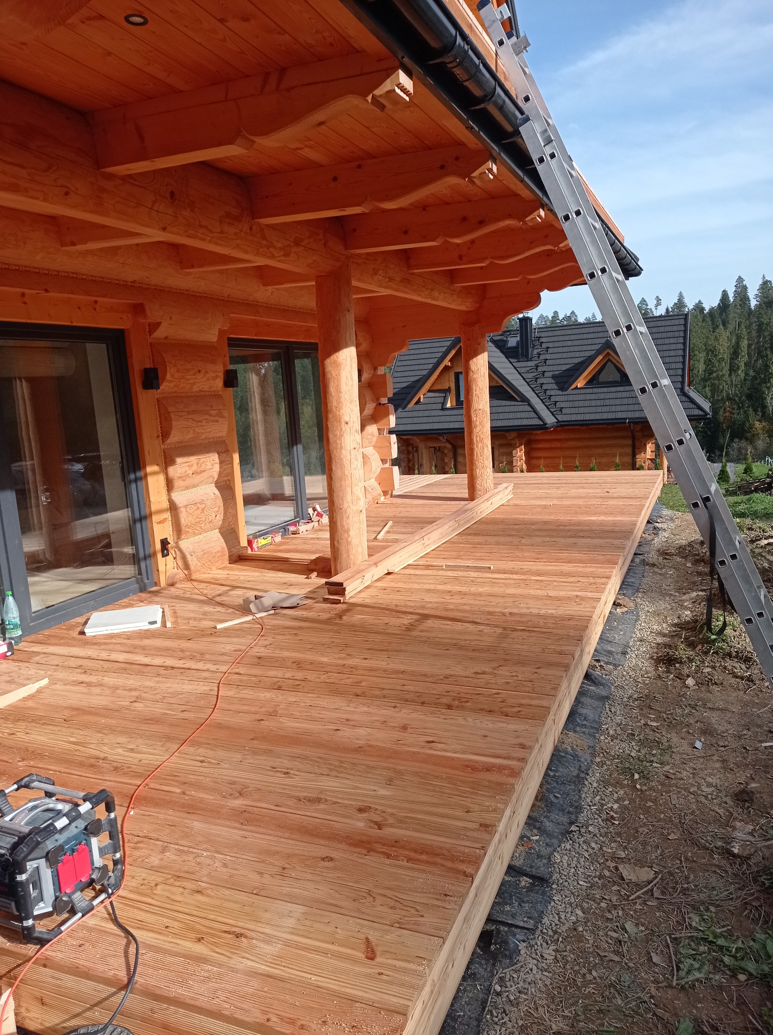 Drewniany taras z bali w trakcie budowy, widoczne narzędzia i drabina oparta o dach, w tle dom z ciemnym dachem.