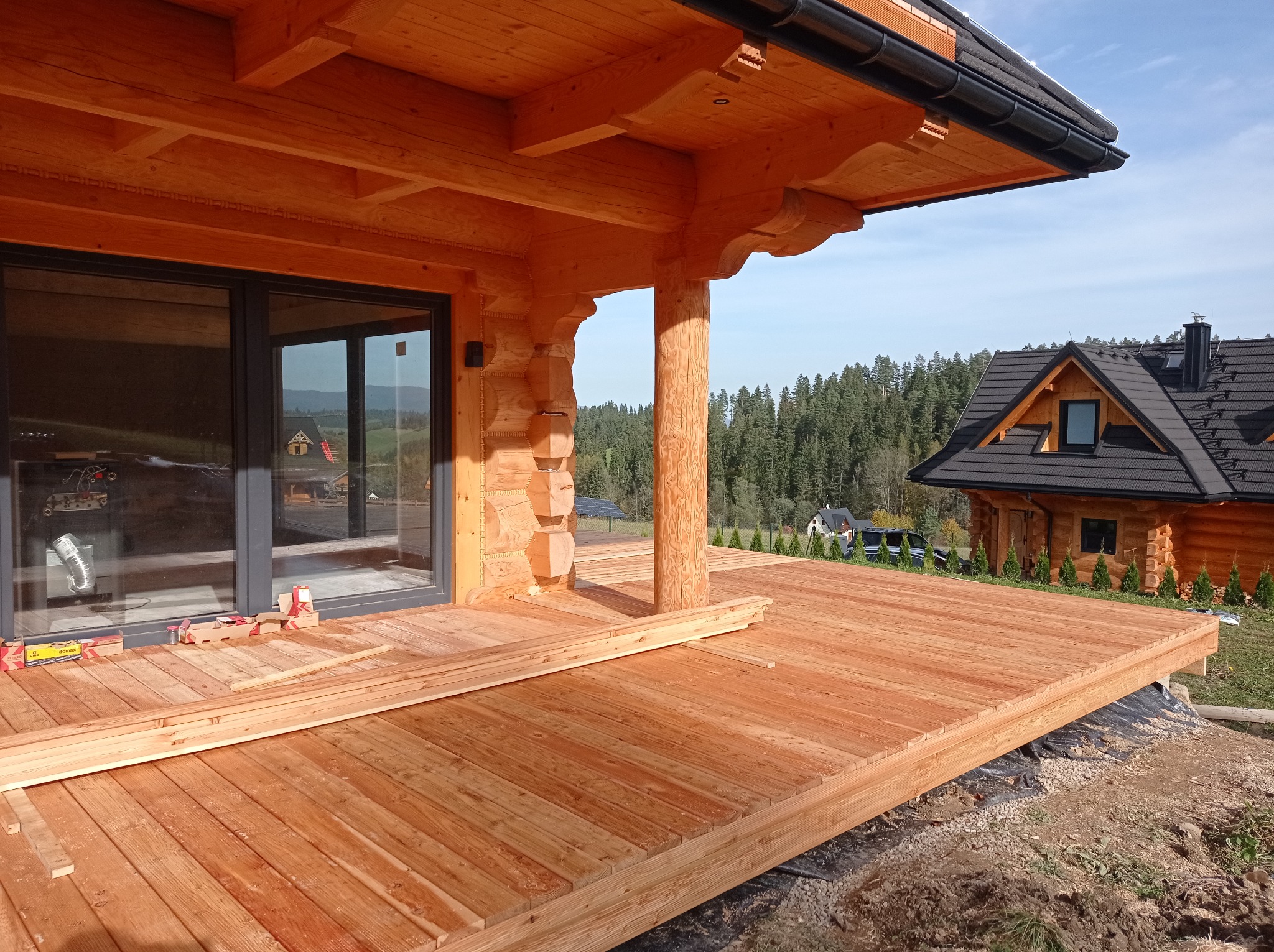 Drewniany taras w trakcie budowy przy domu z bali, widoczne deski podłogowe i materiały budowlane, w tle las i dom z ciemnym dachem.